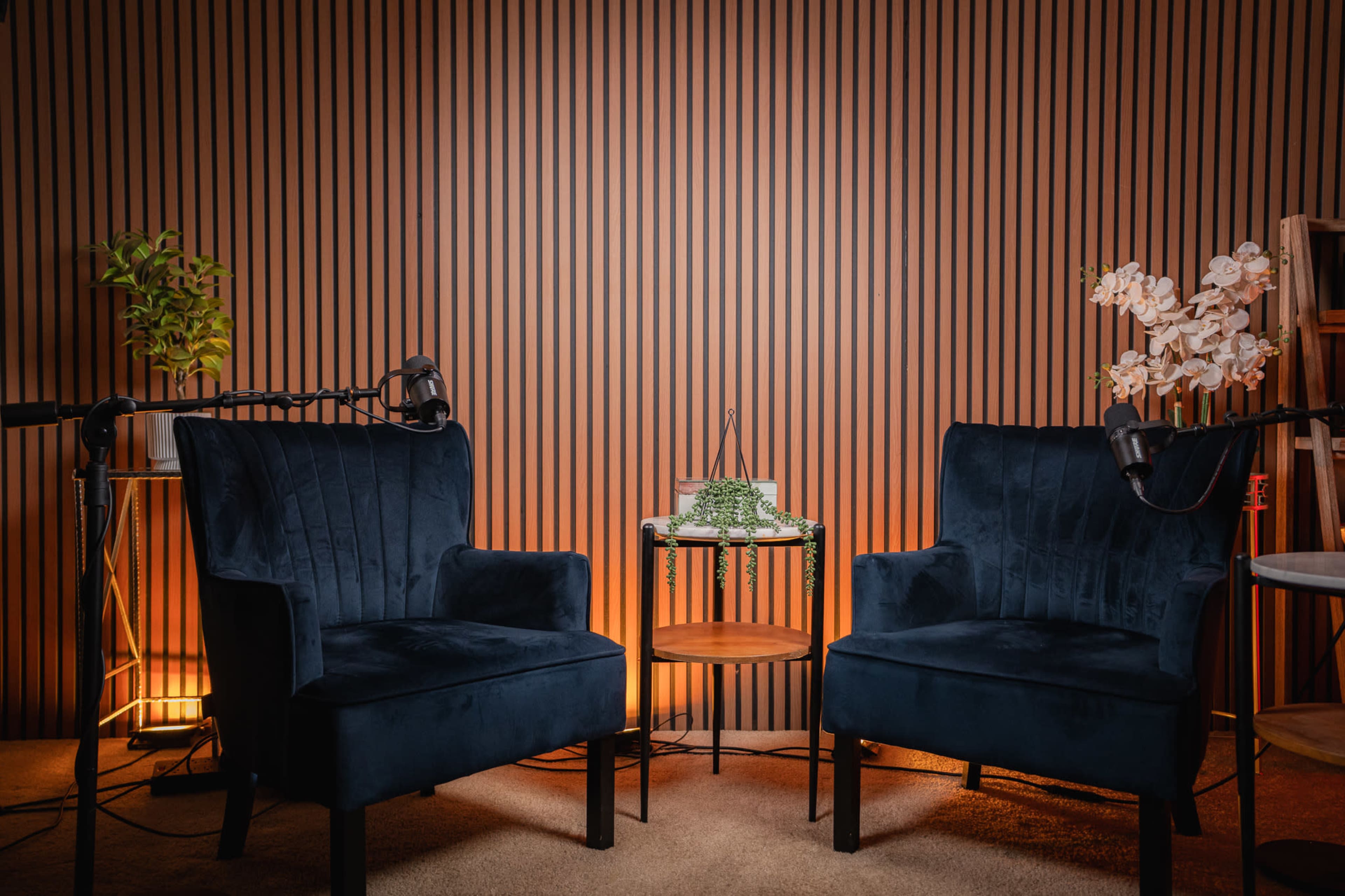 The image shows two blue velvet chairs positioned facing each other, with a small round table between them, all set against a striped wooden wall and soft lighting.
