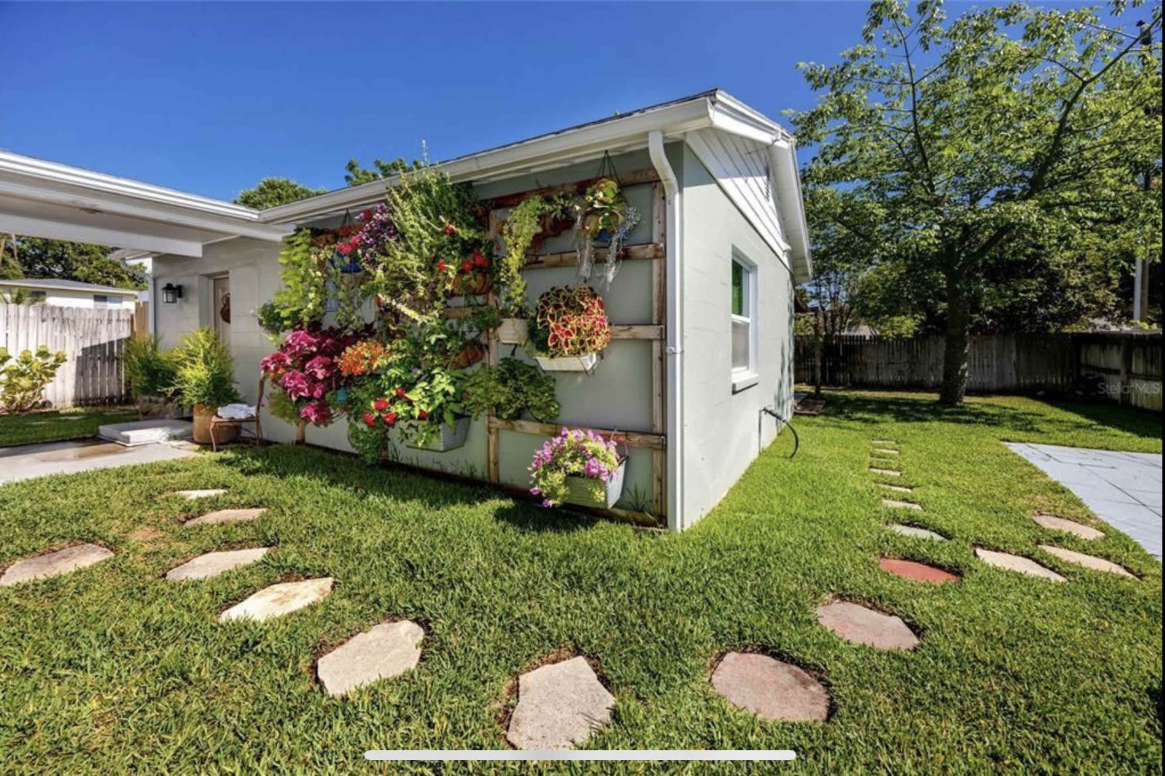 A wall-mounted vertical garden with blooming flowers is displayed beside a gravel path leading to a house, surrounded by green grass.