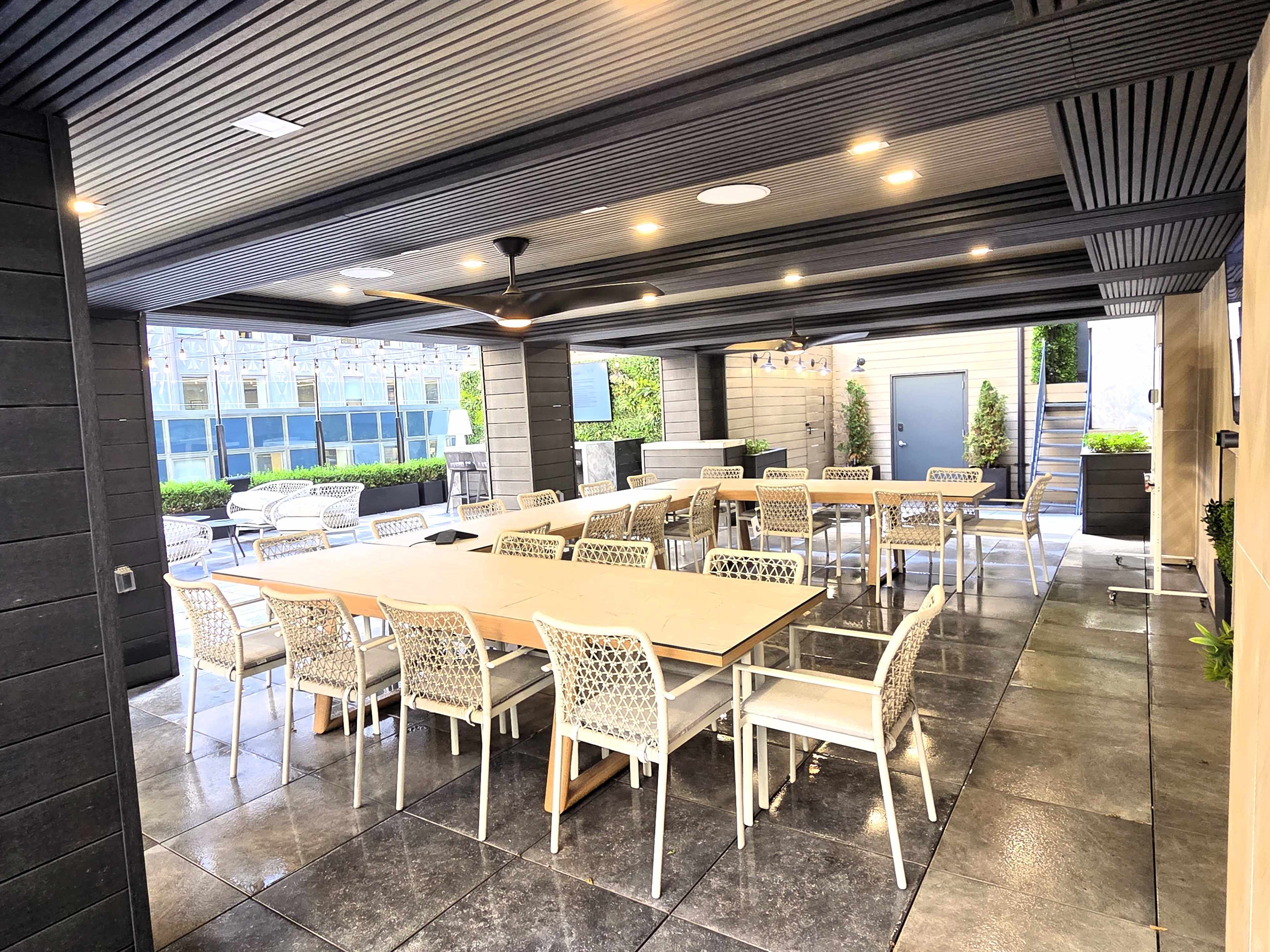 The image shows a spacious outdoor dining area with a long table surrounded by white chairs, set beneath a covered terrace featuring modern lighting and greenery.