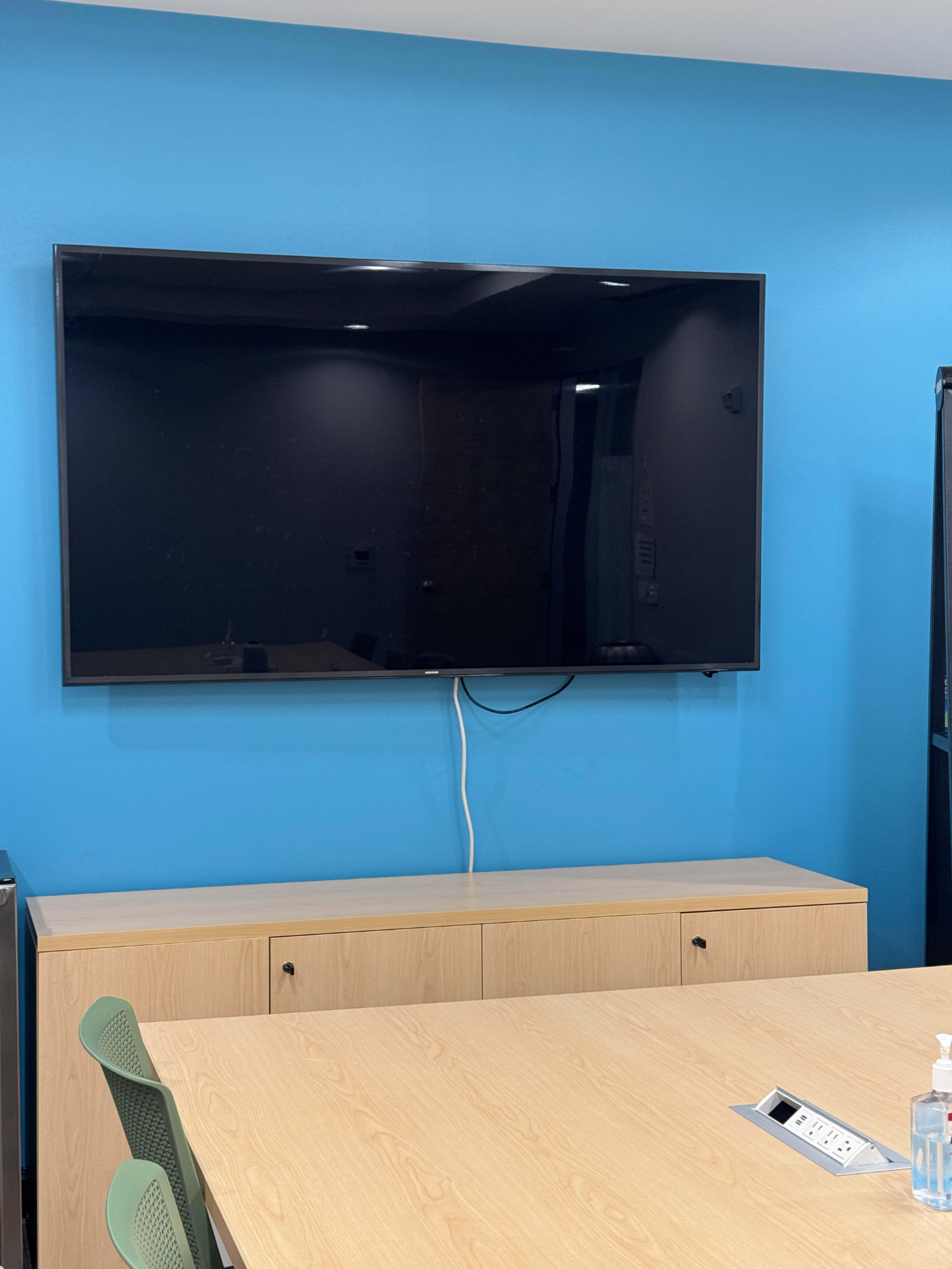 A large flat-screen television is mounted on a blue wall above a light wood credenza in a meeting room.