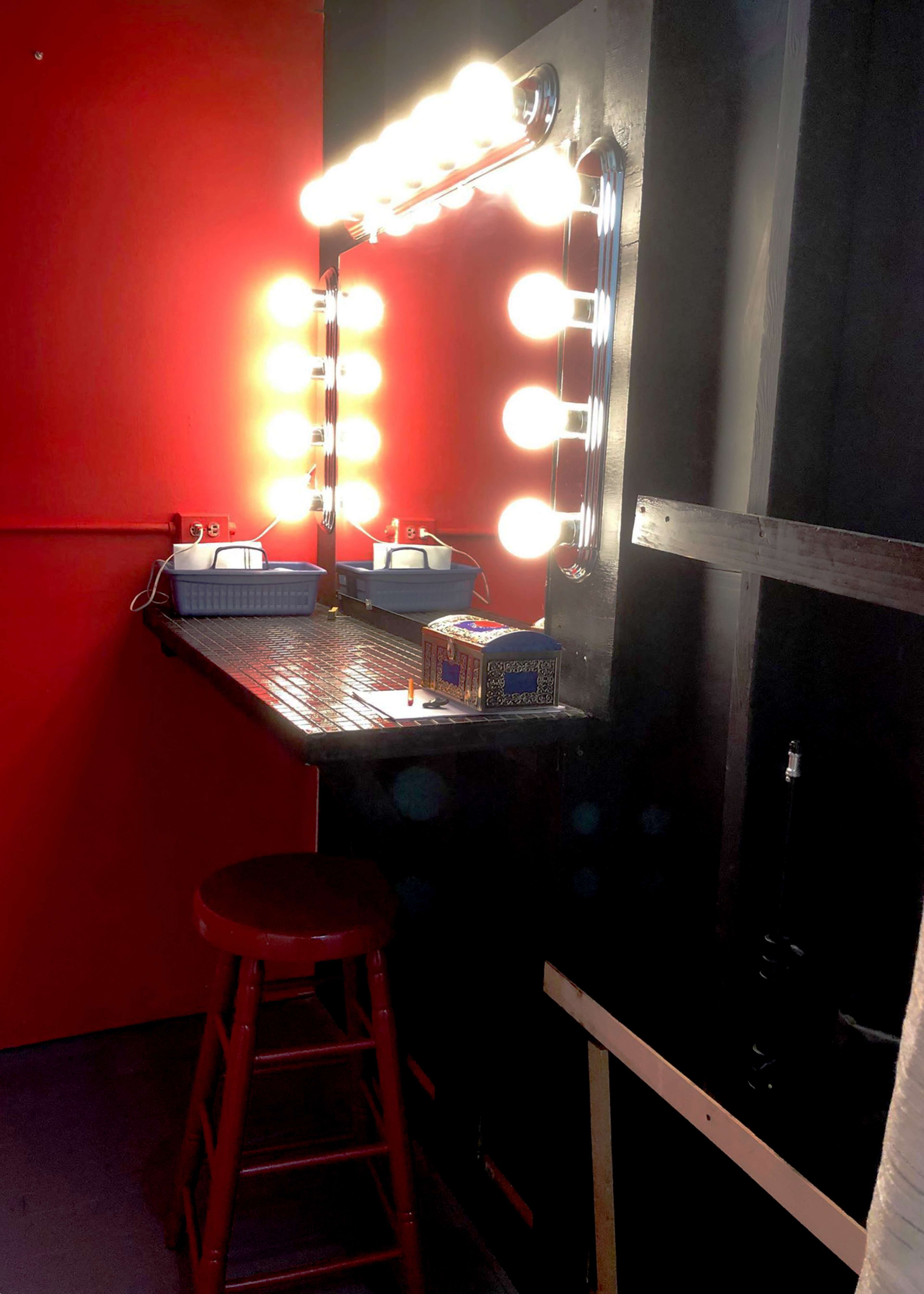 A brightly lit makeup mirror with multiple bulbs sits above a black countertop in a red-walled room.