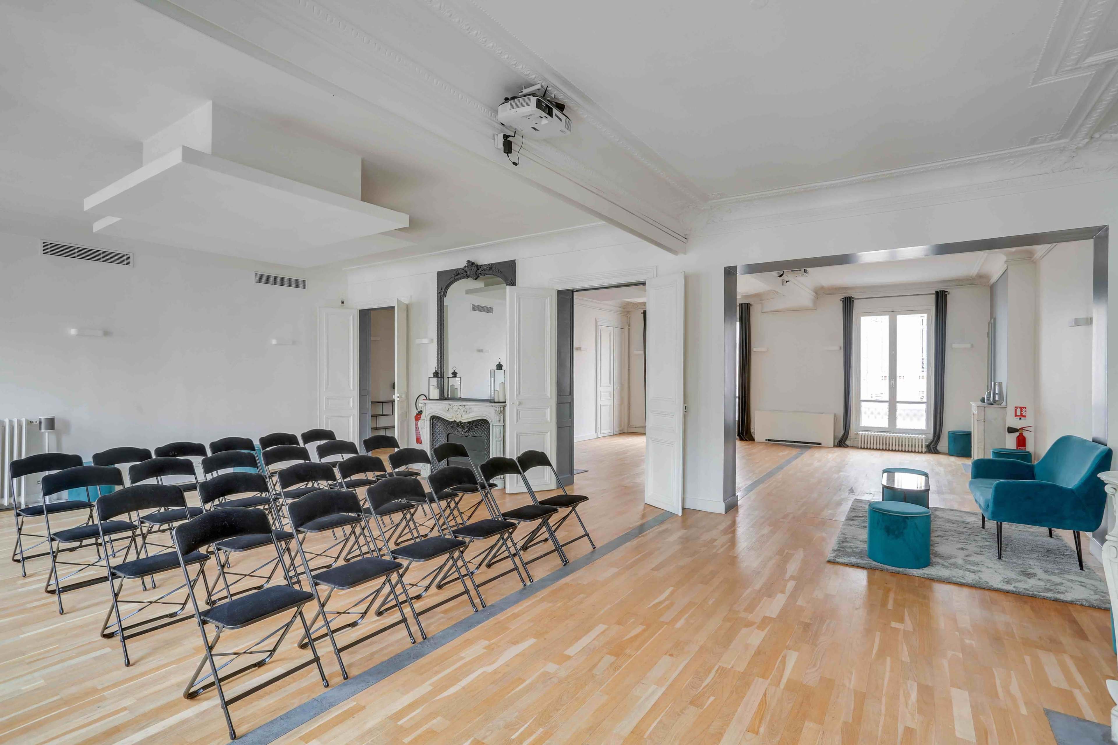A spacious room arranged with rows of black folding chairs, a small seating area in the corner, and large windows allowing natural light.