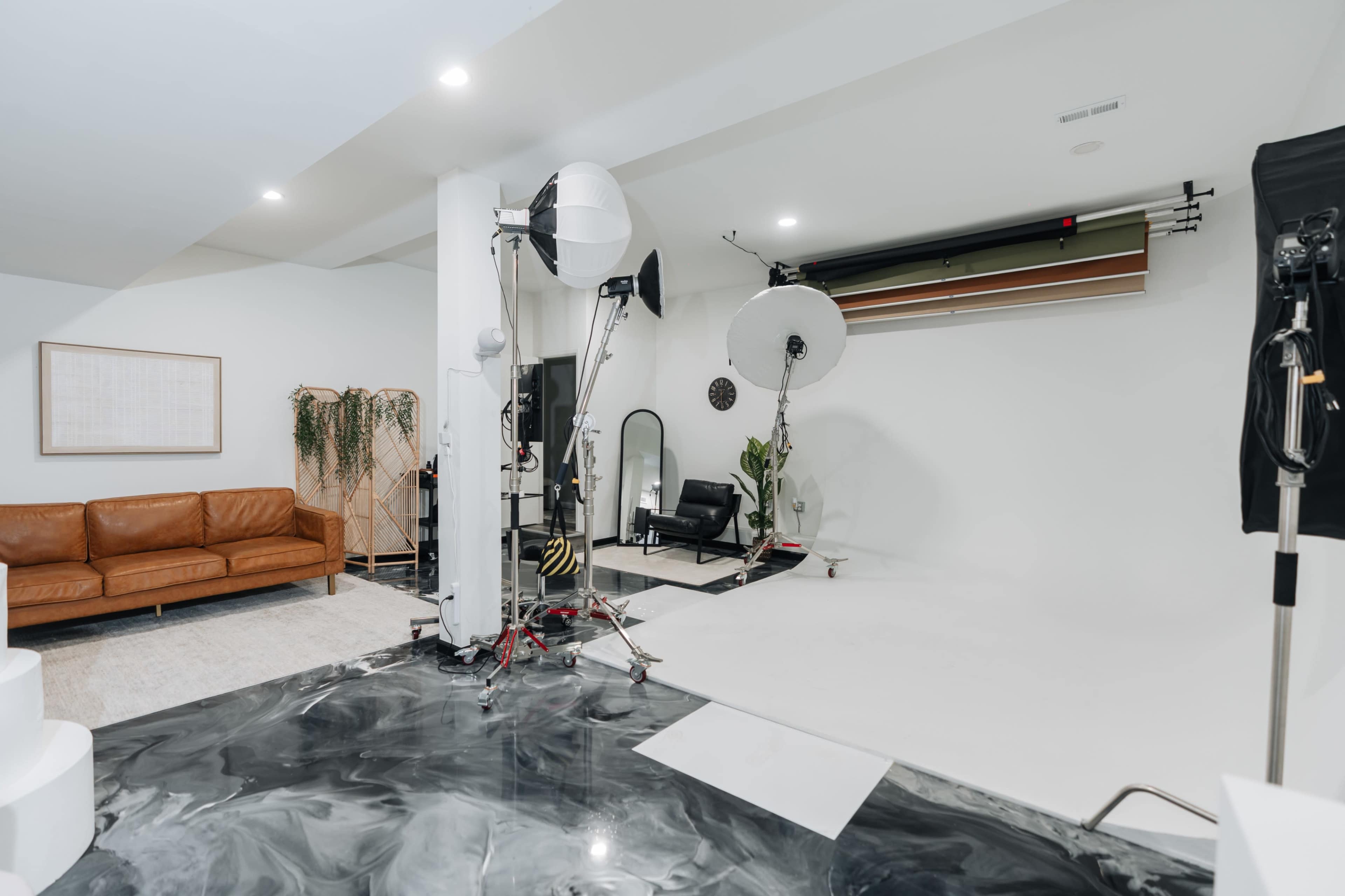The image shows a photography studio with a white backdrop, lighting equipment, and a brown leather couch.