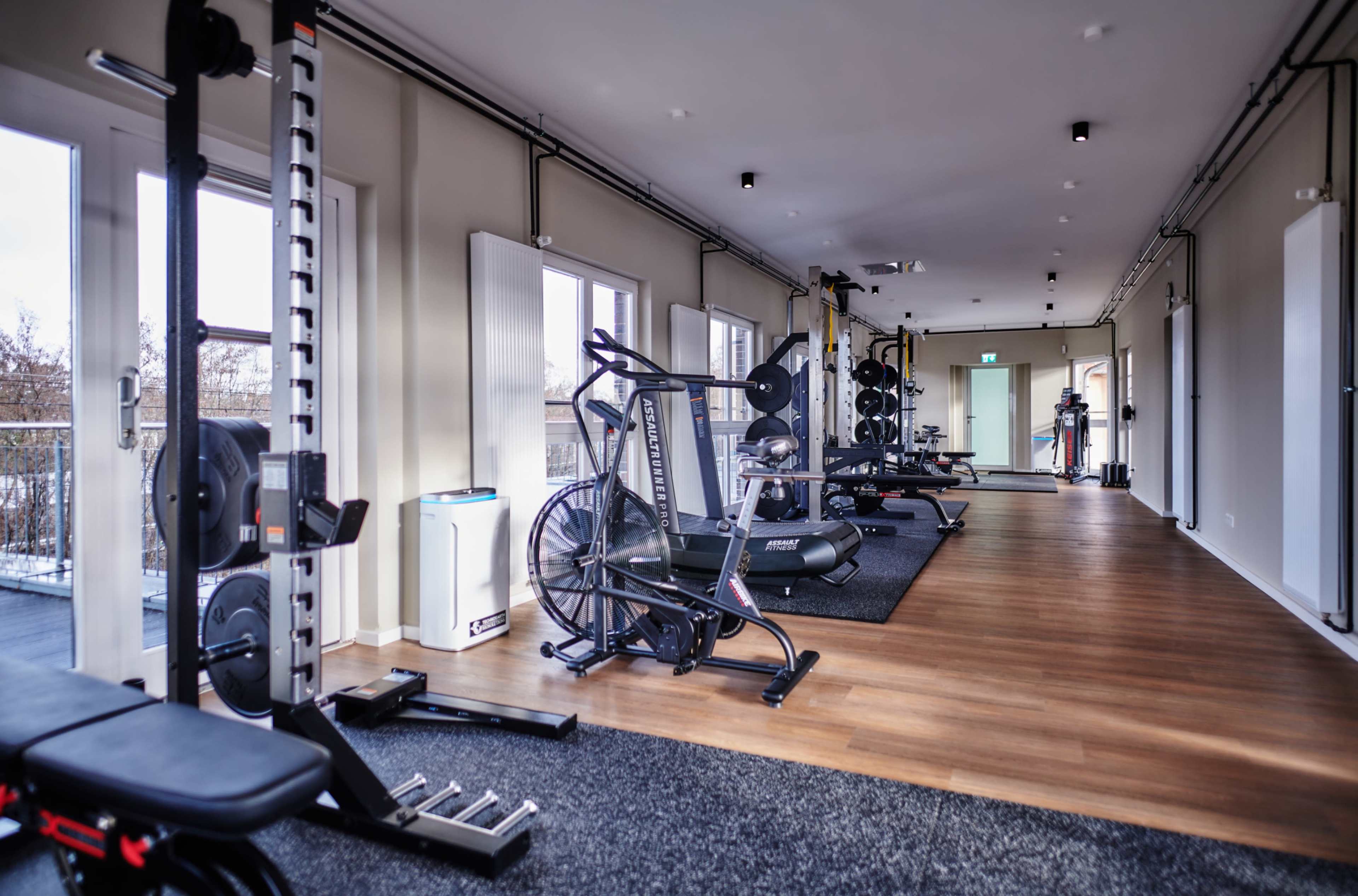 The image shows a modern gym interior with various fitness equipment arranged along a long, well-lit room featuring windows and wooden flooring.