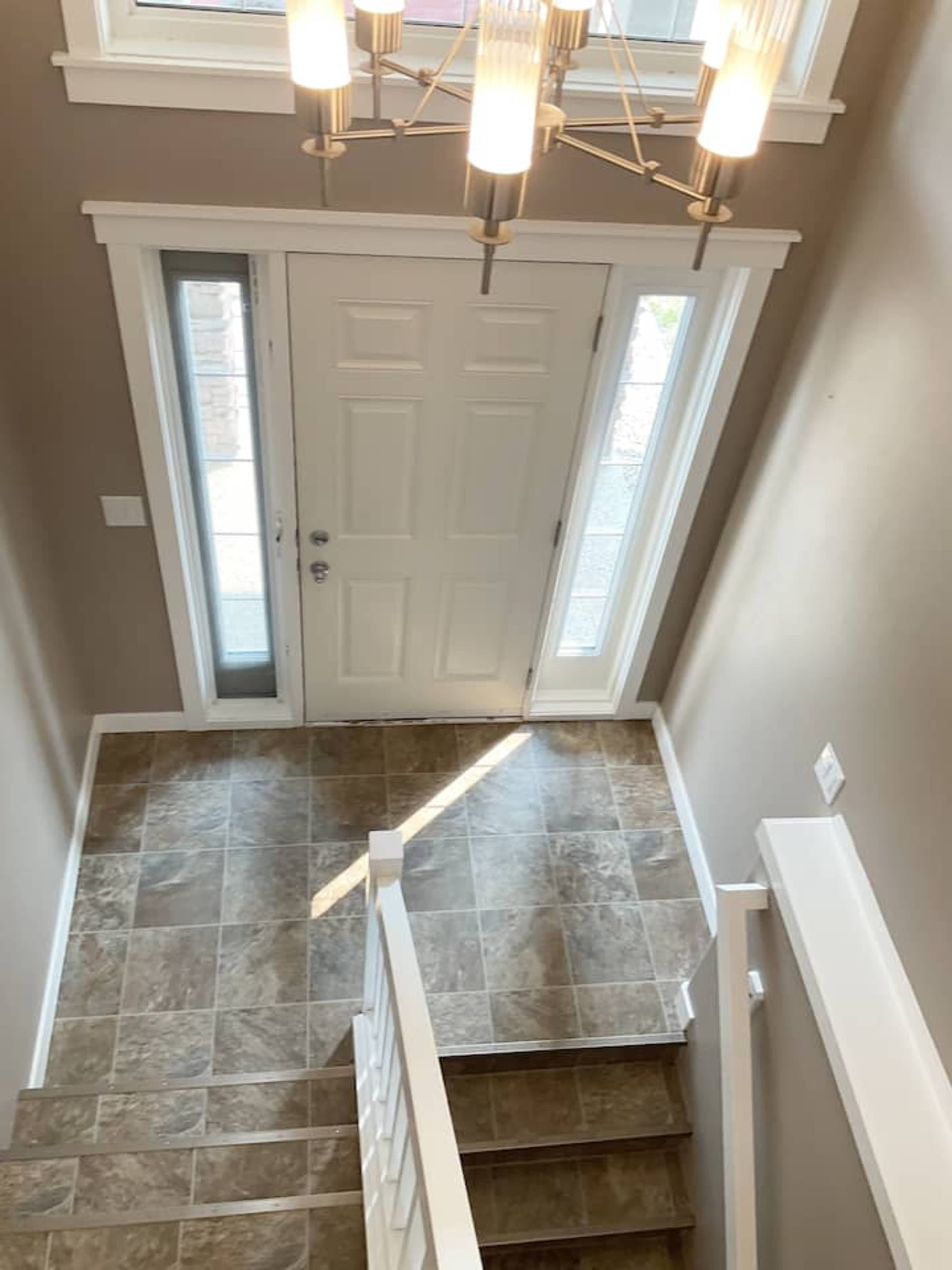 A well-lit entrance hall with a staircase leading down, a front door with two sidelights, and tiled flooring.