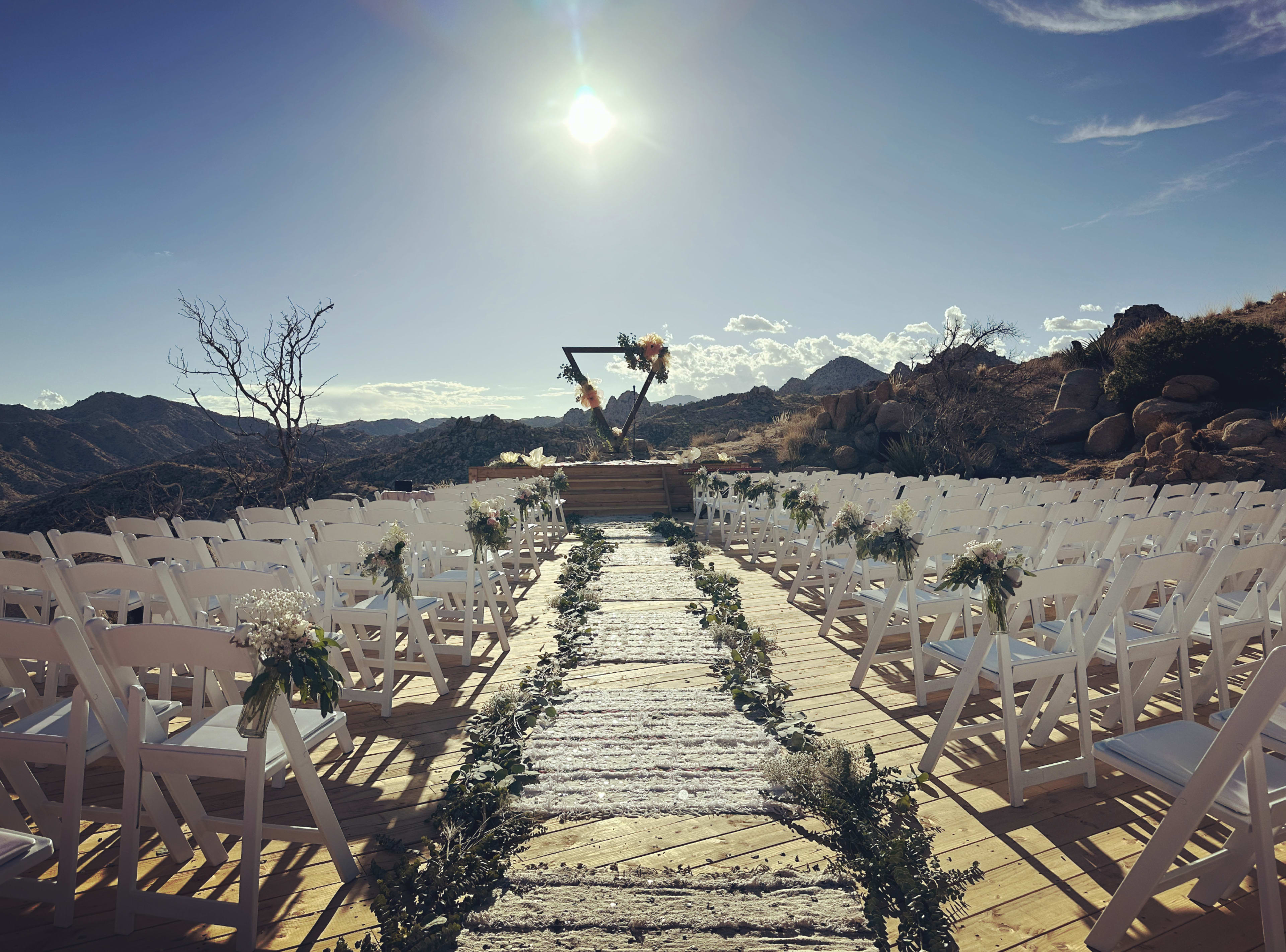 A scenic outdoor wedding venue features rows of white chairs facing an altar adorned with flowers, set against a mountainous backdrop under a bright sun.