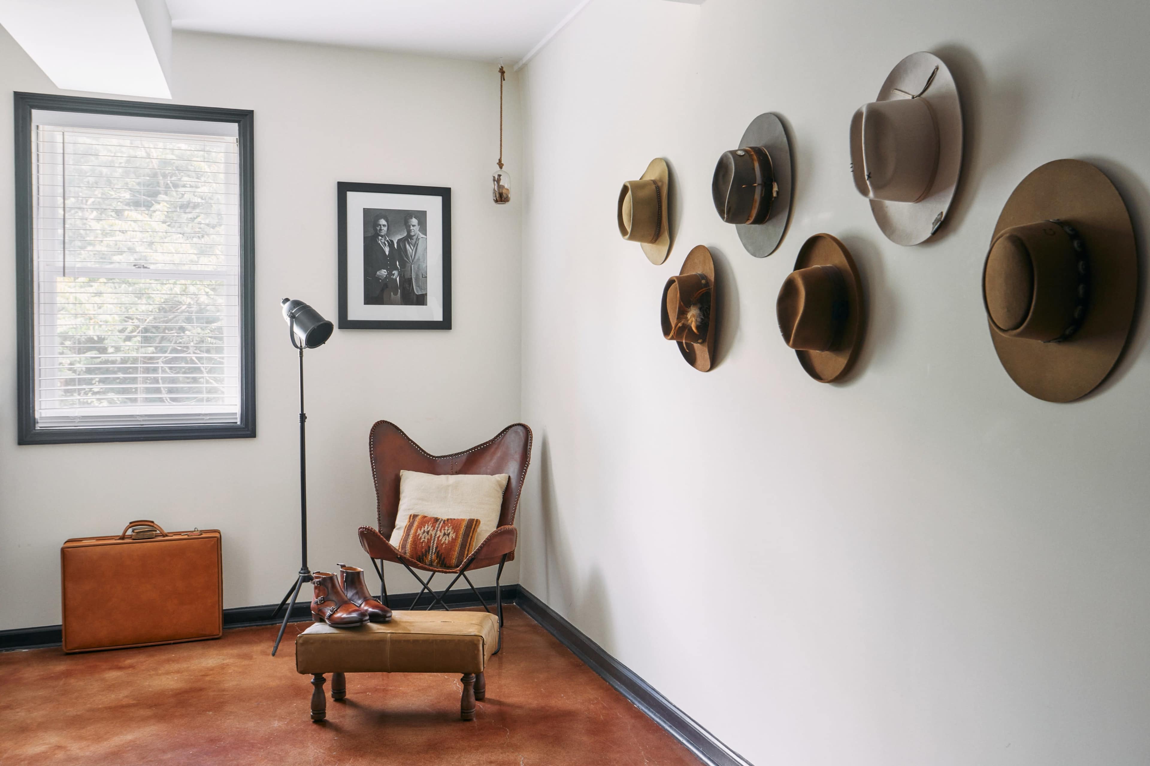 A cozy room featuring a butterfly chair, a vintage suitcase, and a wall adorned with various hats, along with a framed photo and a floor lamp.