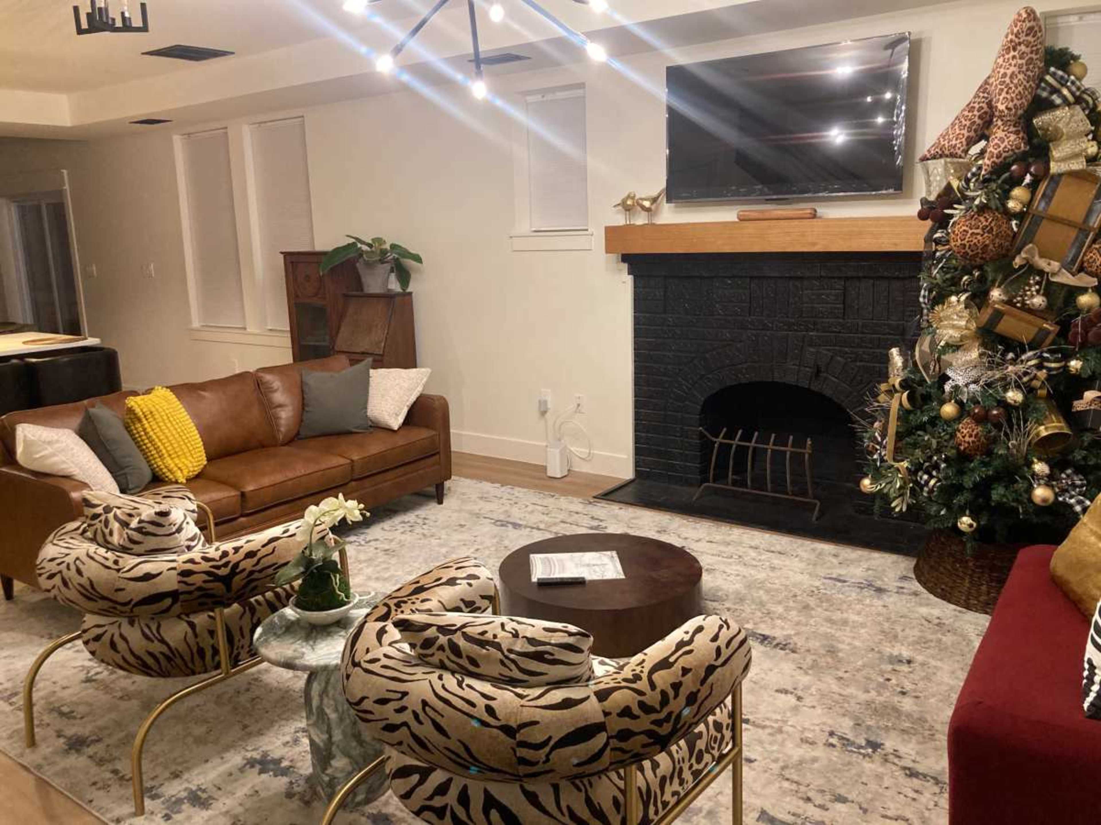 The image shows a stylish living room featuring a large Christmas tree, a comfortable brown leather sofa, two patterned chairs, and a modern fireplace with a television above it.
