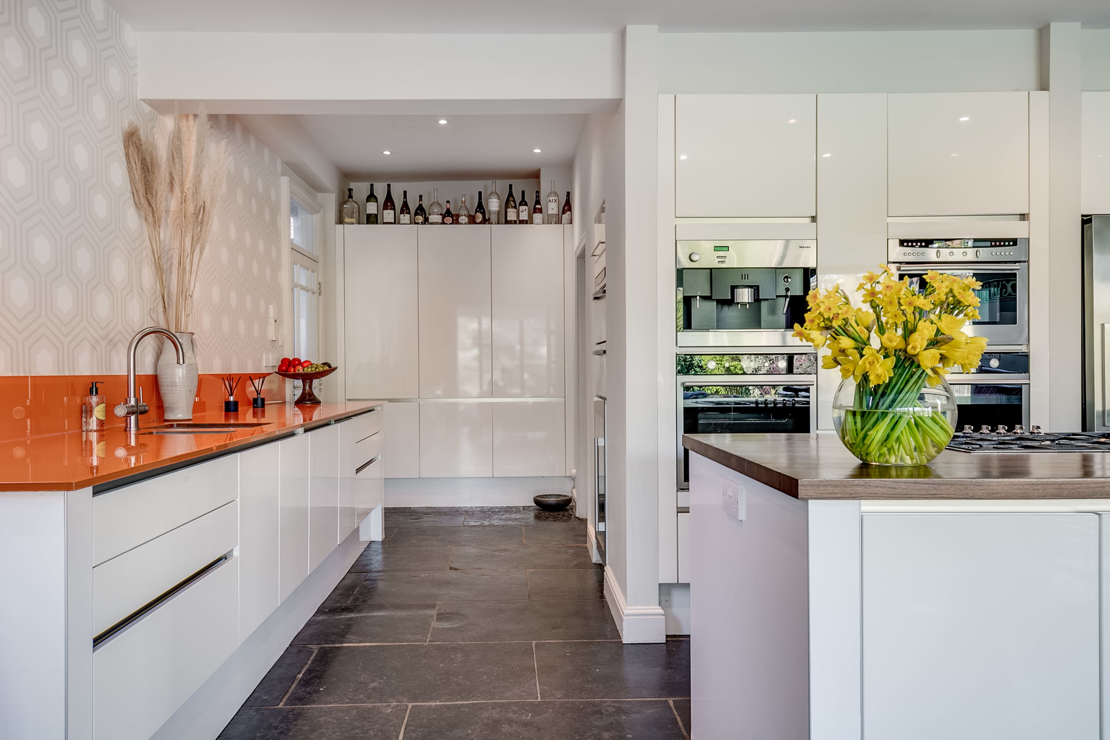 The kitchen features sleek white cabinetry, an orange backsplash, and a vase of yellow flowers on a wooden island.