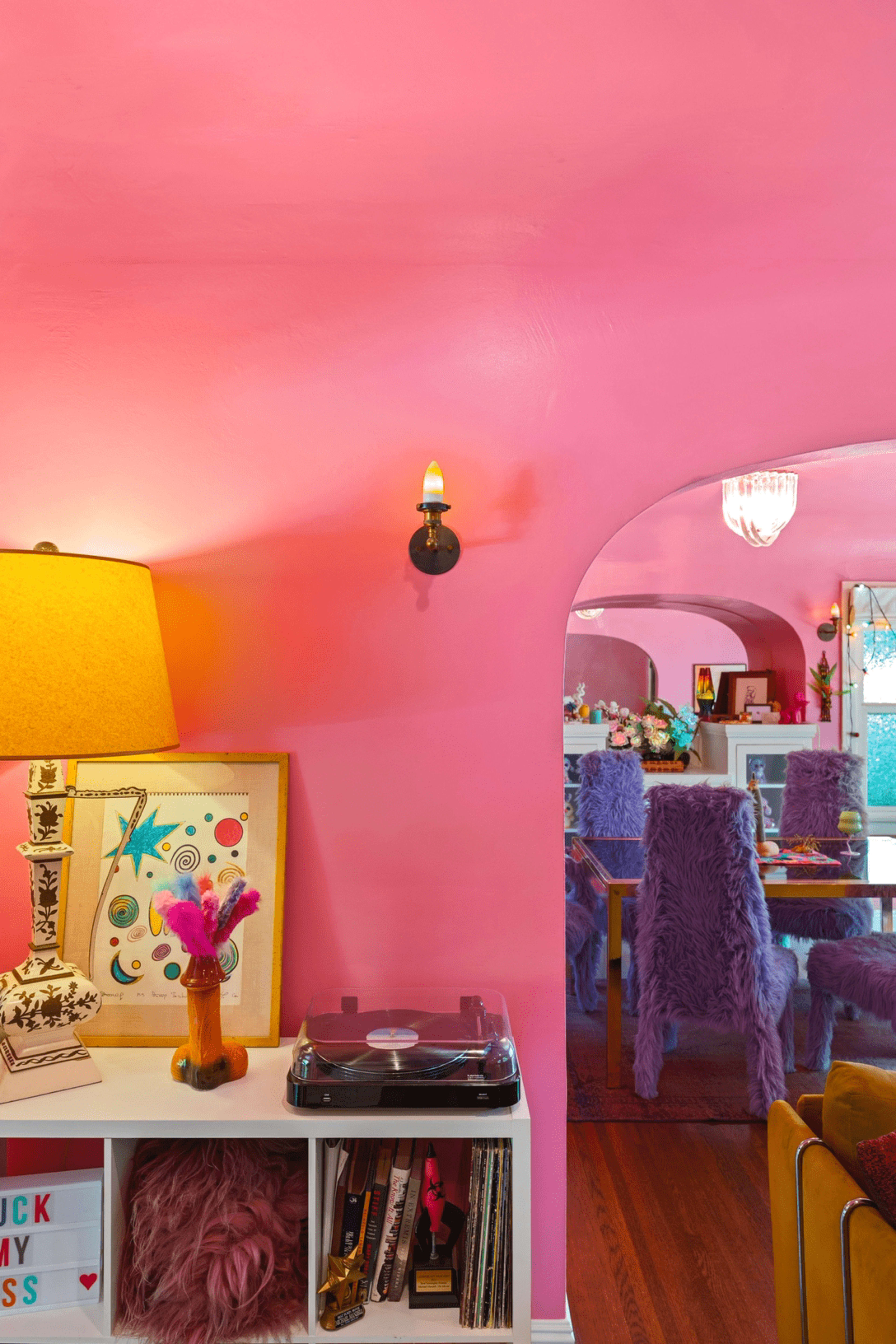 The image shows a brightly colored pink room featuring a lamp, a record player on a shelf, and a dining area with fuzzy chairs in the background.
