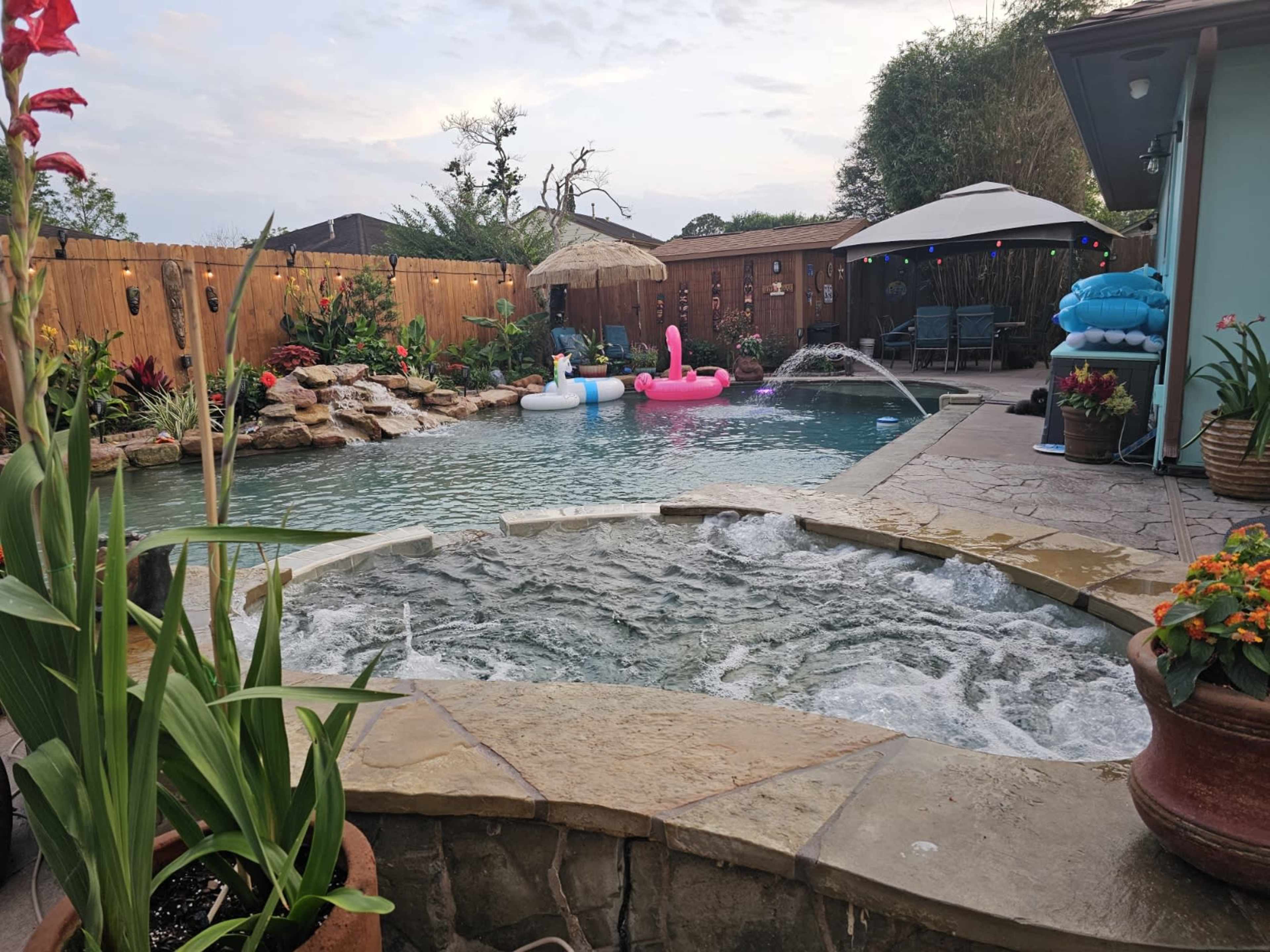 A backyard pool area featuring a jacuzzi, two inflatable flamingos, and lounge chairs under a shaded umbrella, surrounded by decorative plants and a stone patio.