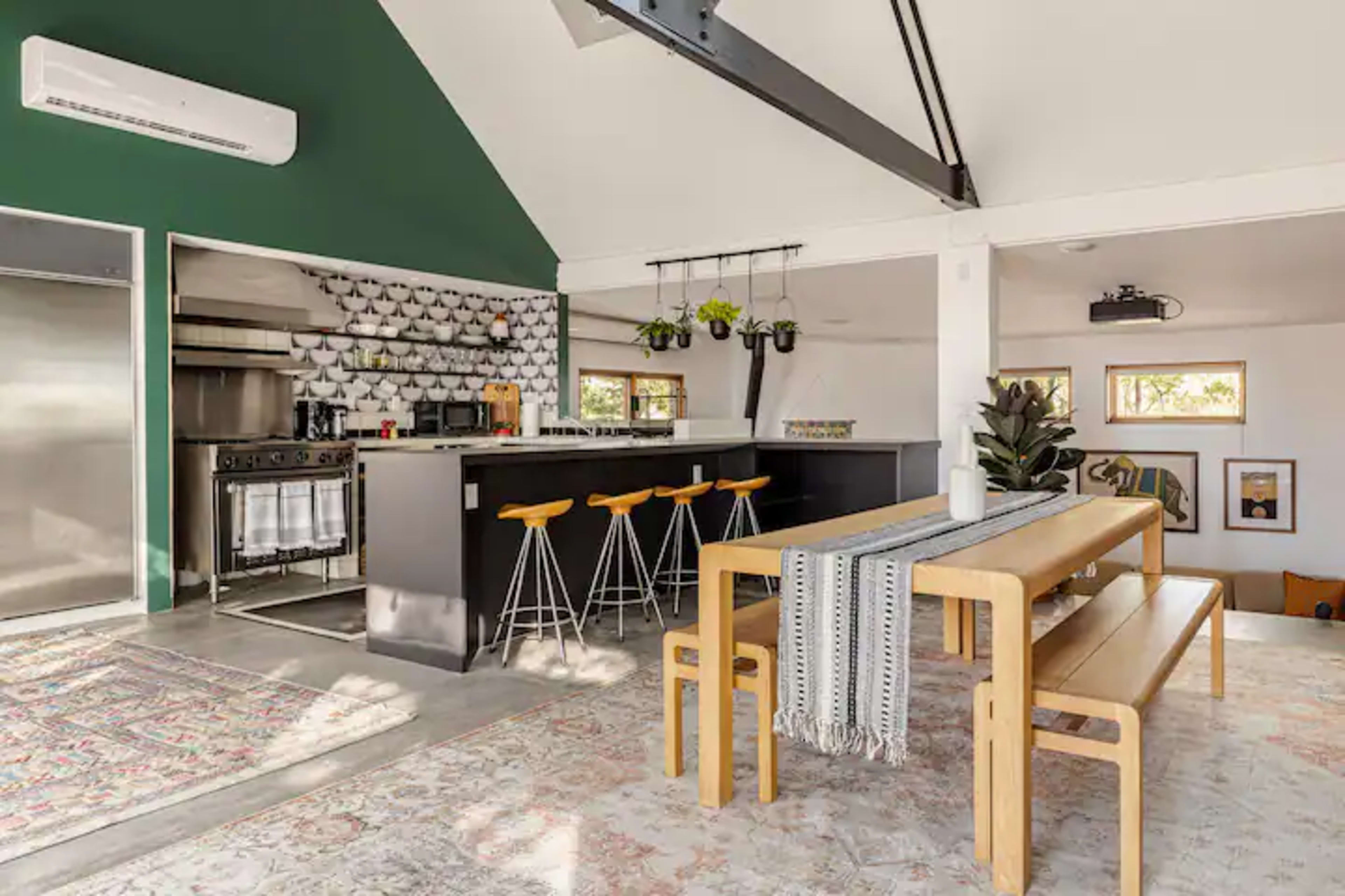 The image shows a modern kitchen and dining area featuring a dark green accent wall, a large kitchen island with bar stools, and a long wooden dining table set on a patterned rug.