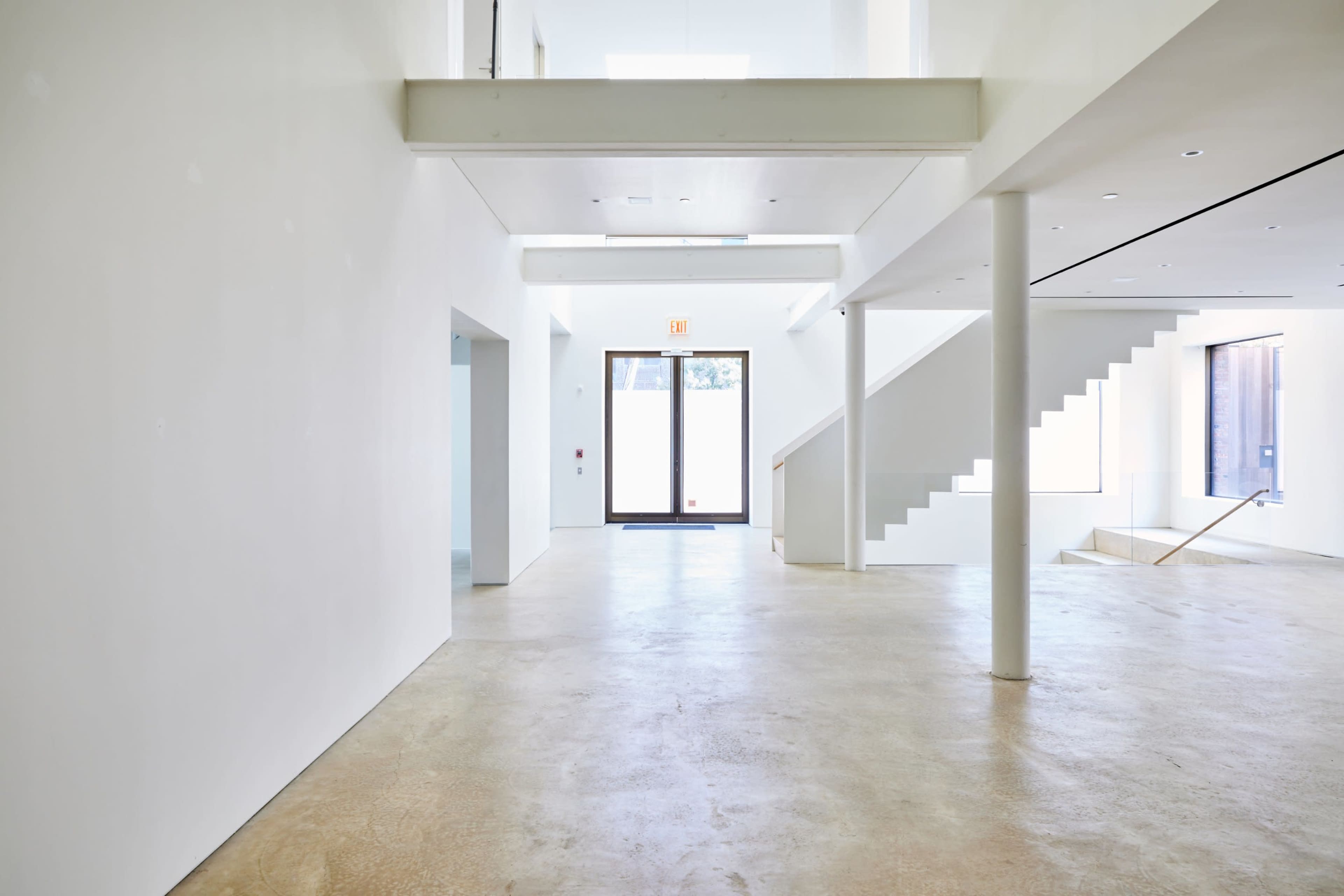 The image shows a spacious, modern interior with white walls, a concrete floor, and a staircase leading to an upper level.