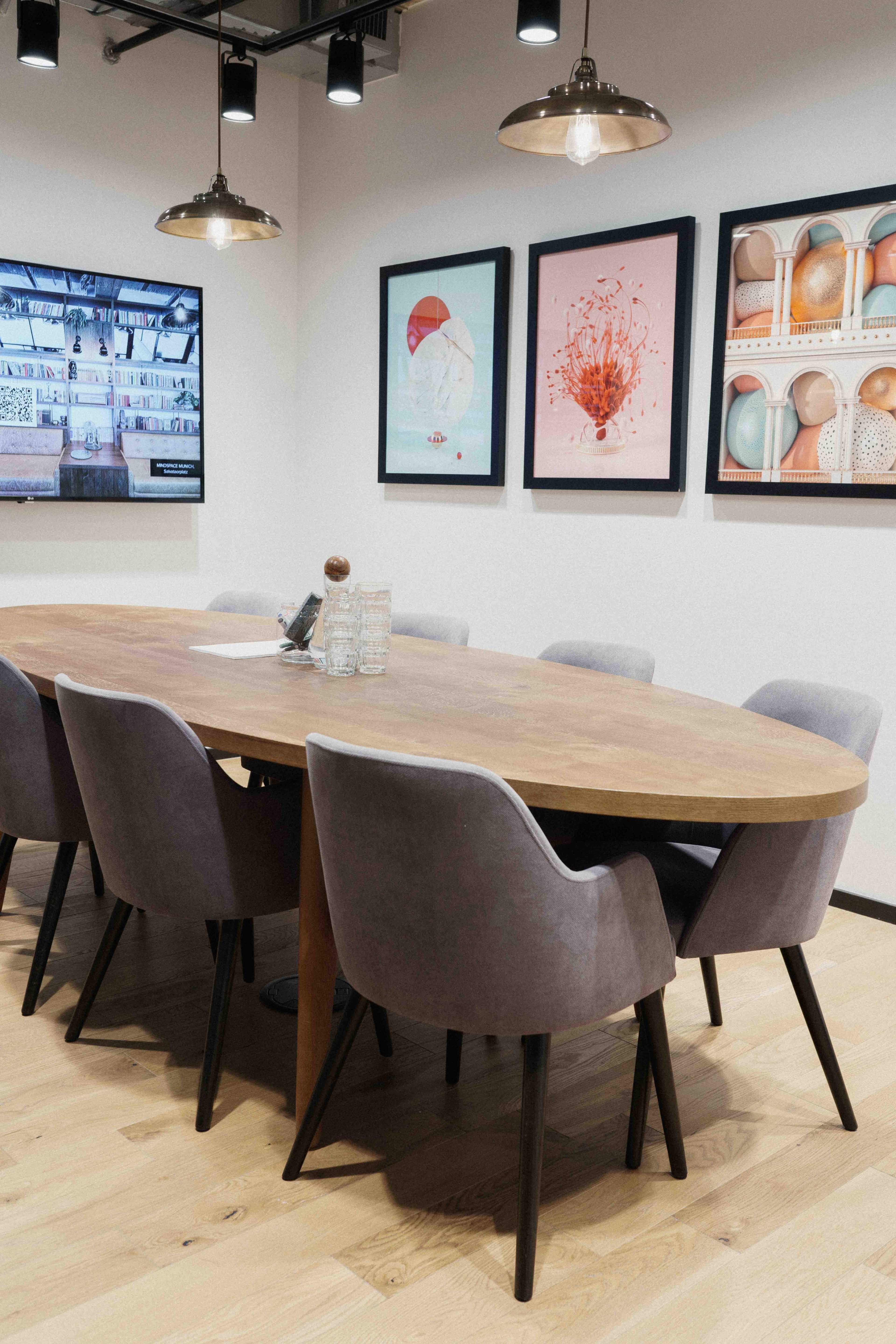 A large, oval wooden conference table is surrounded by gray upholstered chairs in a modern meeting room featuring framed artwork on the walls.