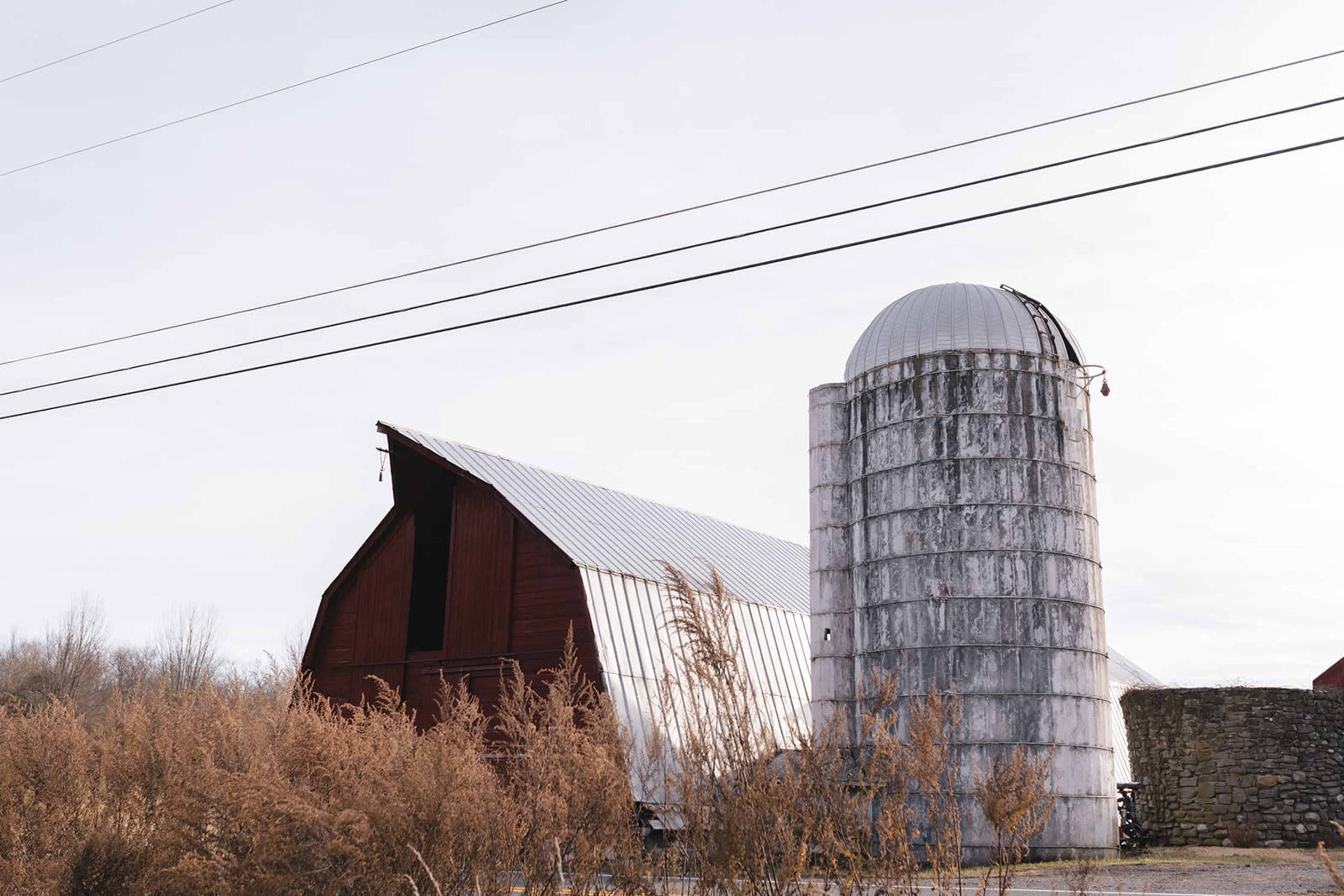 A red barn with a metal roof and a gray silo stands beside a field of tall grass under a cloudy sky.