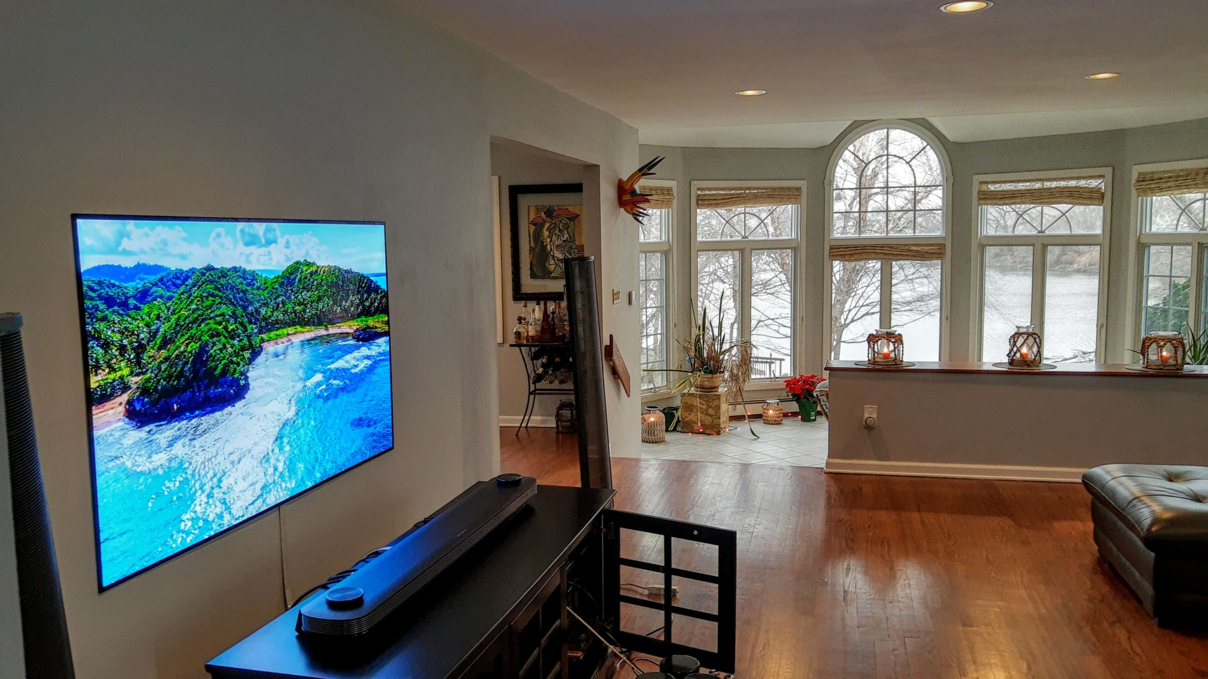 A large television displaying a beach scene is mounted on a wall in a living room with large windows overlooking a body of water.