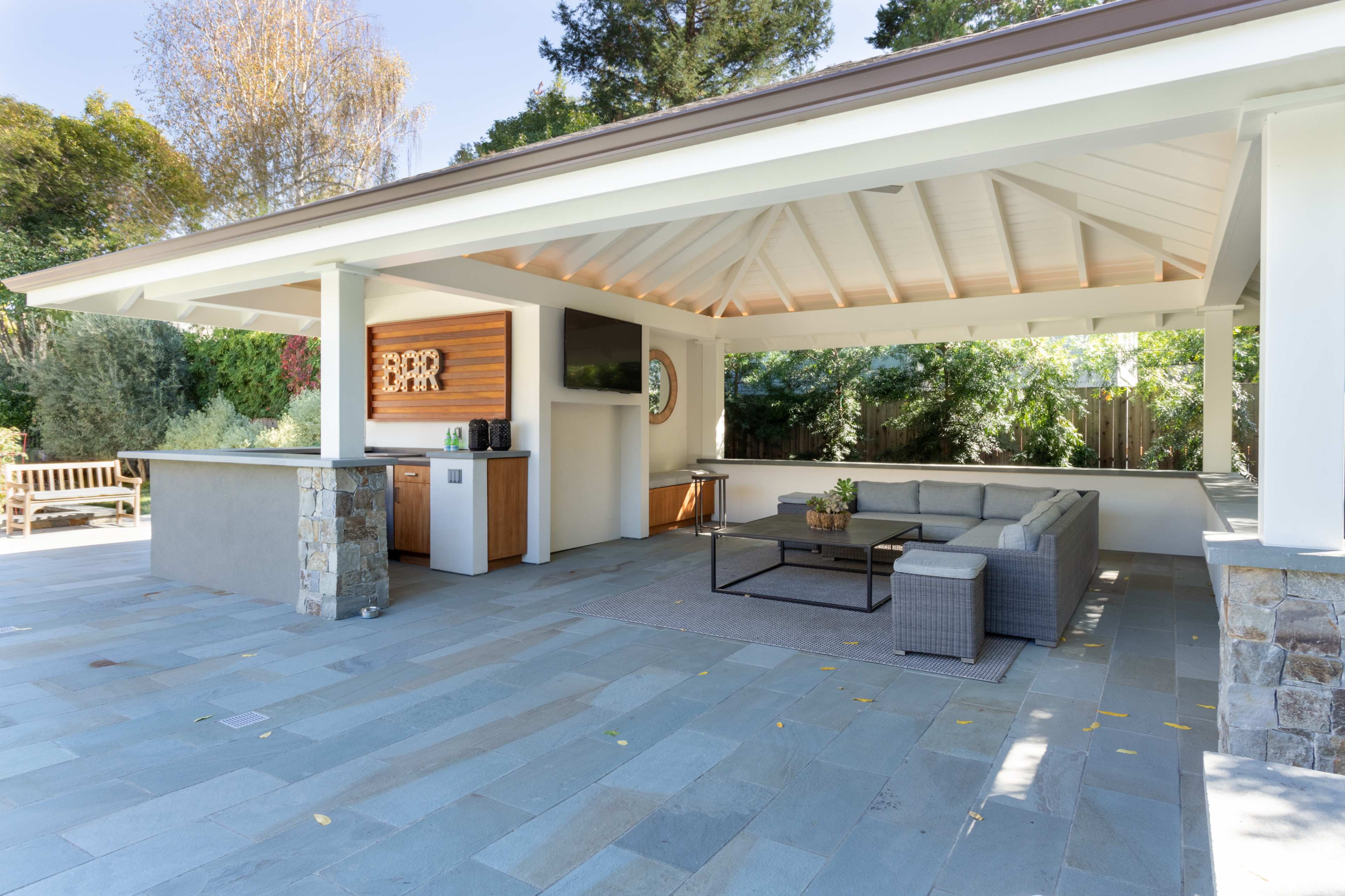 A covered outdoor seating area with a stone bar, gray sectional sofa, and a large flat-screen TV in a landscaped backyard.