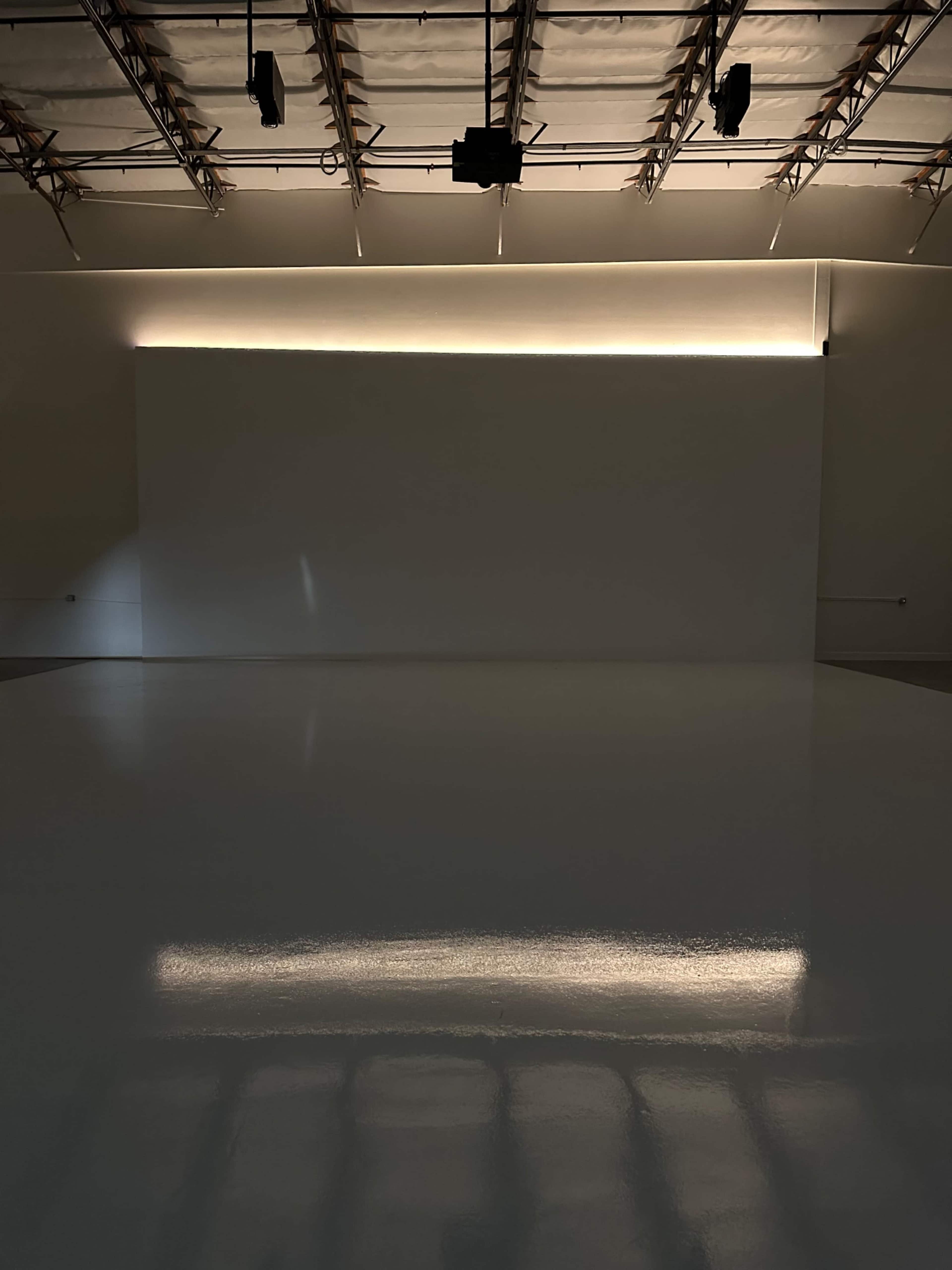 The image shows a minimalist studio space with a glossy white floor and soft lighting illuminating a blank wall.