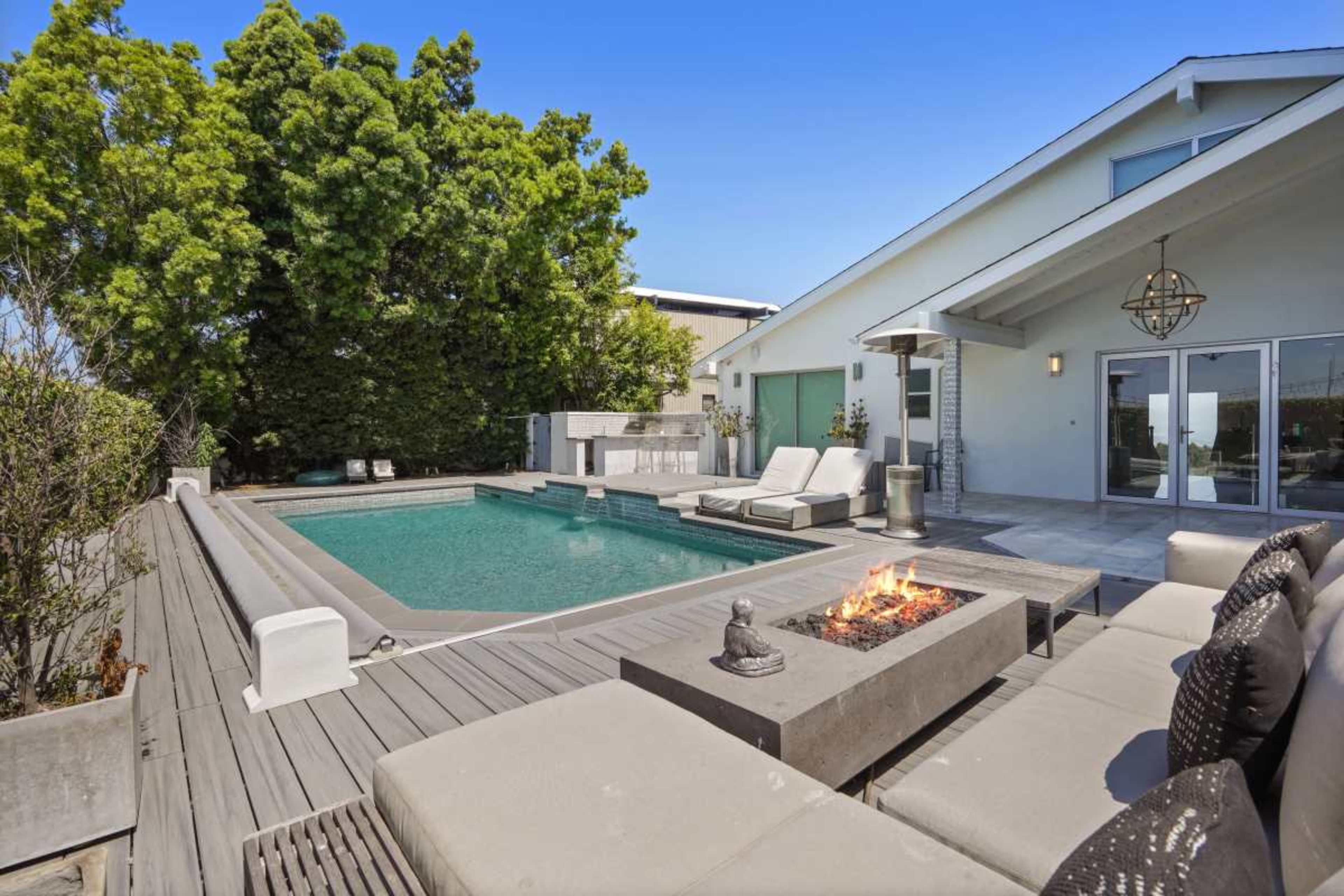 The image shows a backyard featuring a rectangular swimming pool surrounded by lounge chairs, a fire pit, and greenery.