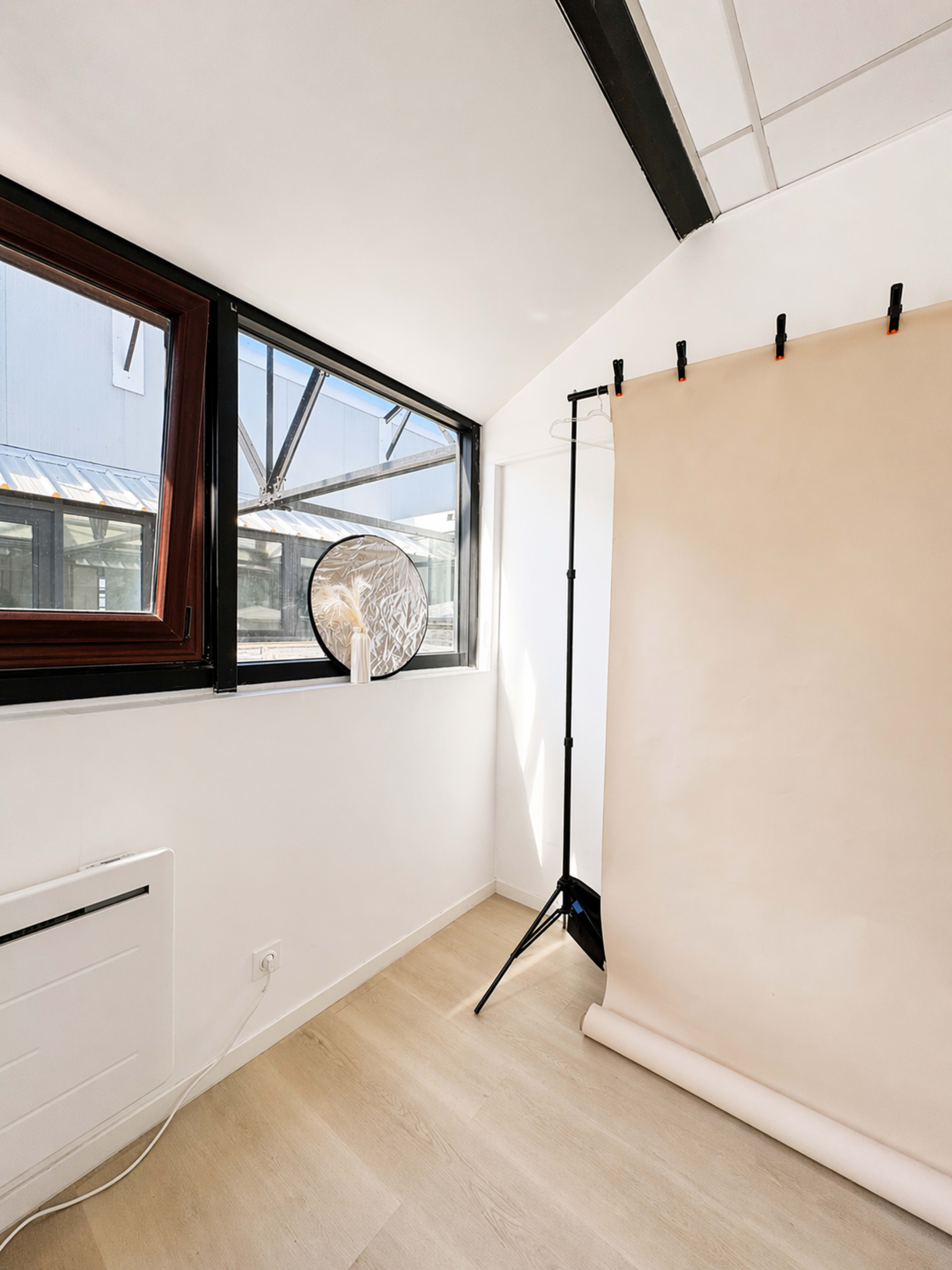 The image shows a bright studio space with a large window, a round reflector, and a backdrop stand holding a beige backdrop.