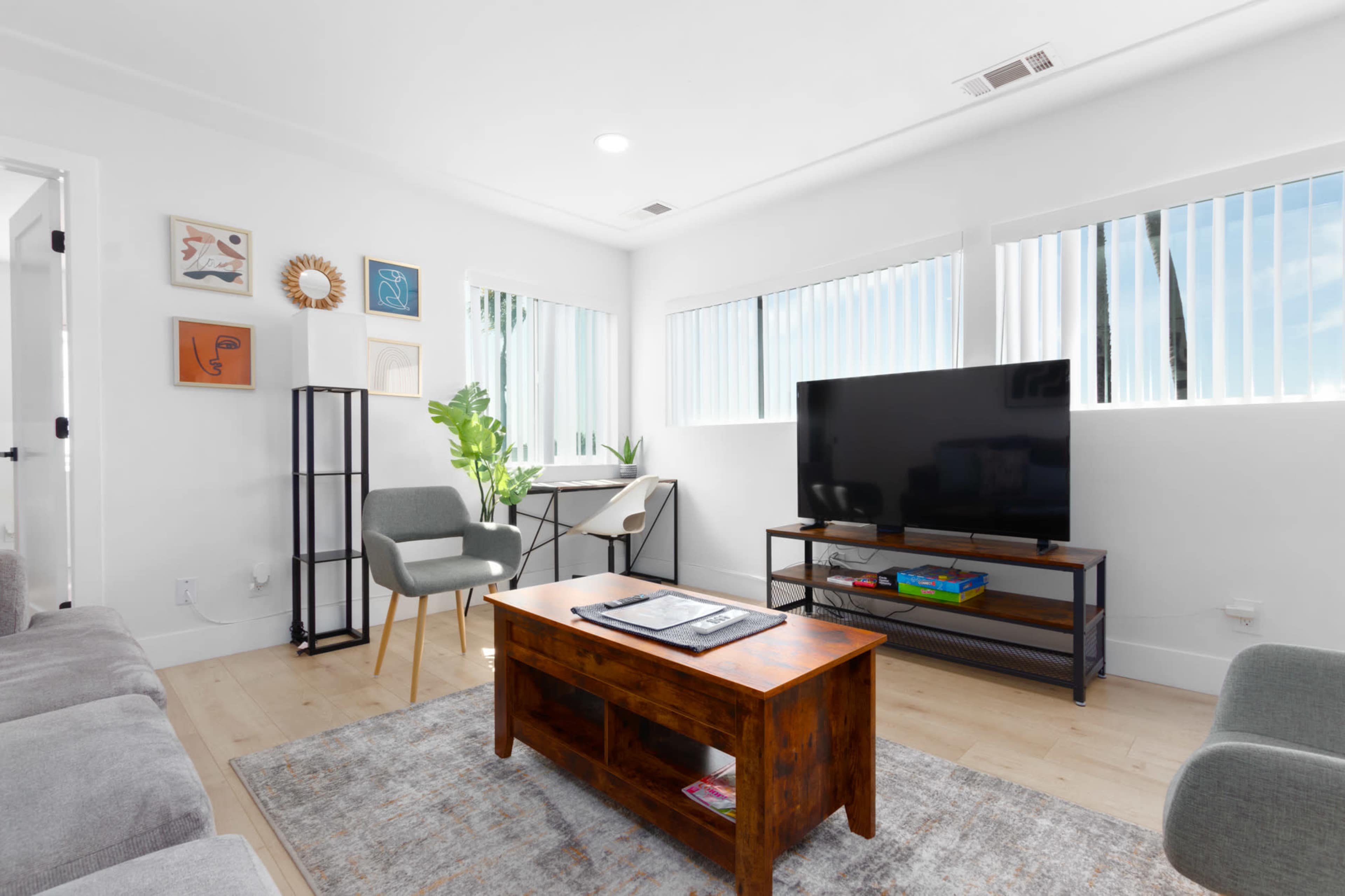 A modern living room features a wooden coffee table, a gray sofa, a television on a metal stand, and large windows allowing natural light.