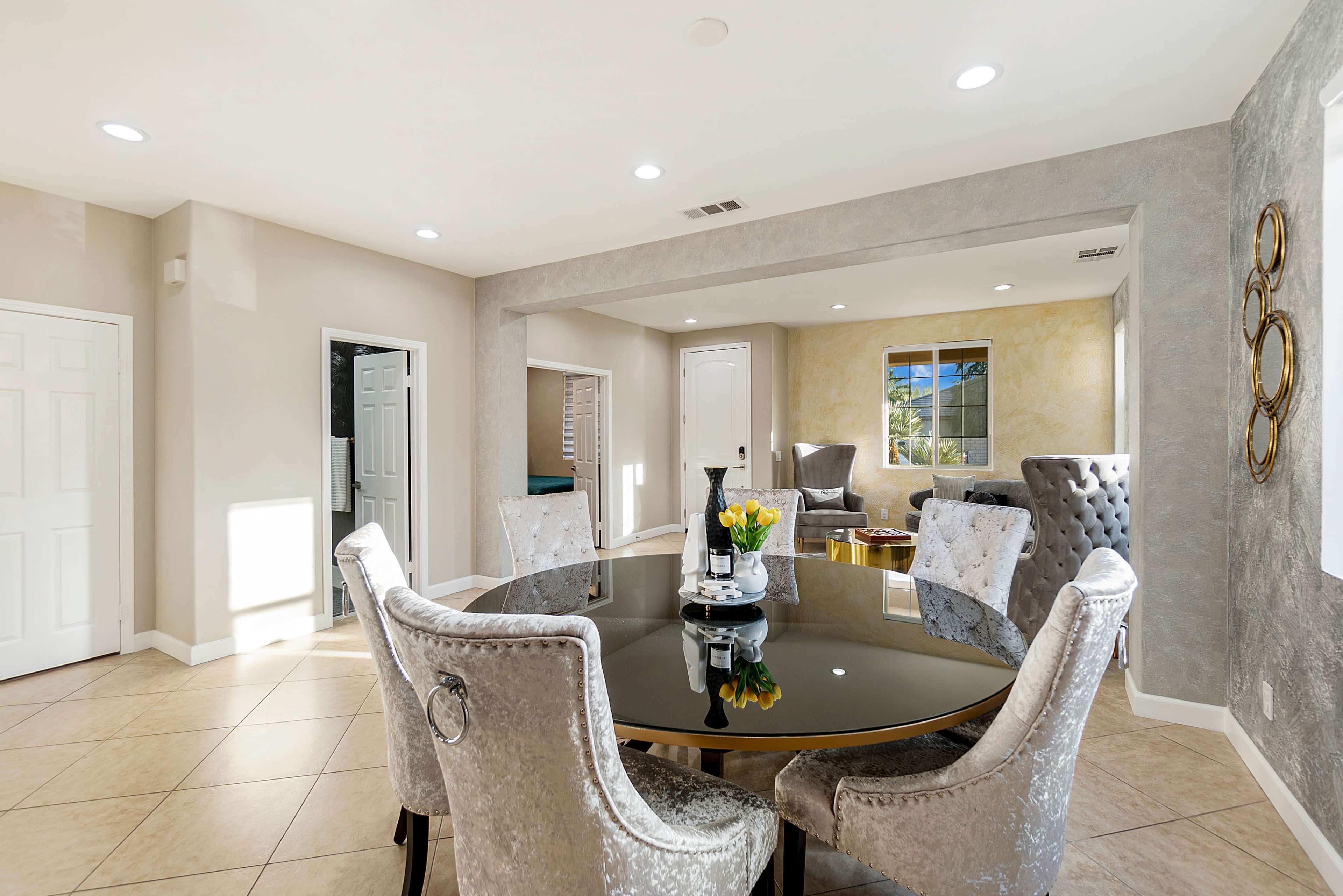 A dining area features a round black table surrounded by silver upholstered chairs, with natural light coming in through large windows.