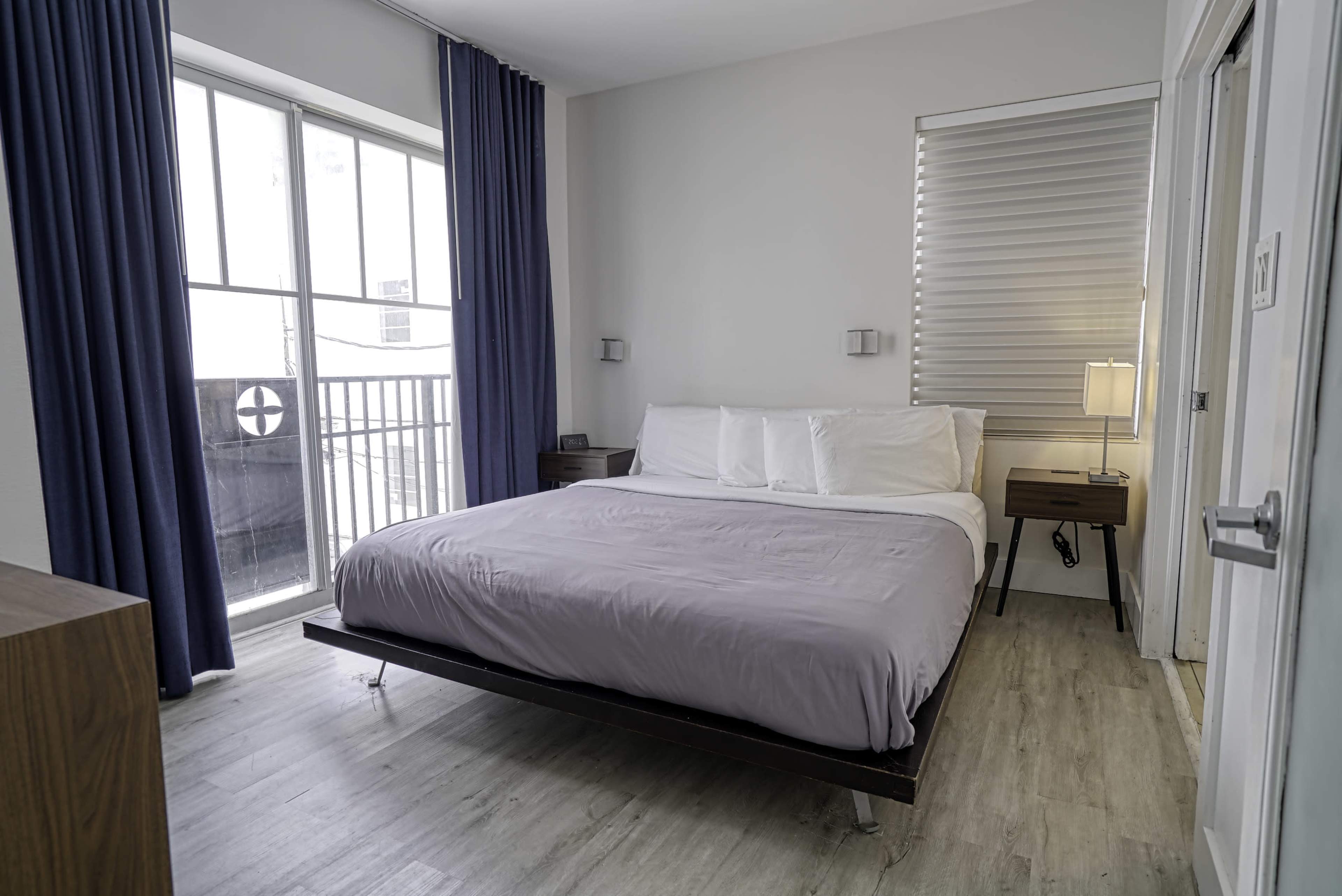 The image shows a modern bedroom with a large bed, dark wood nightstands, and blue curtains, featuring a window that overlooks a balcony.