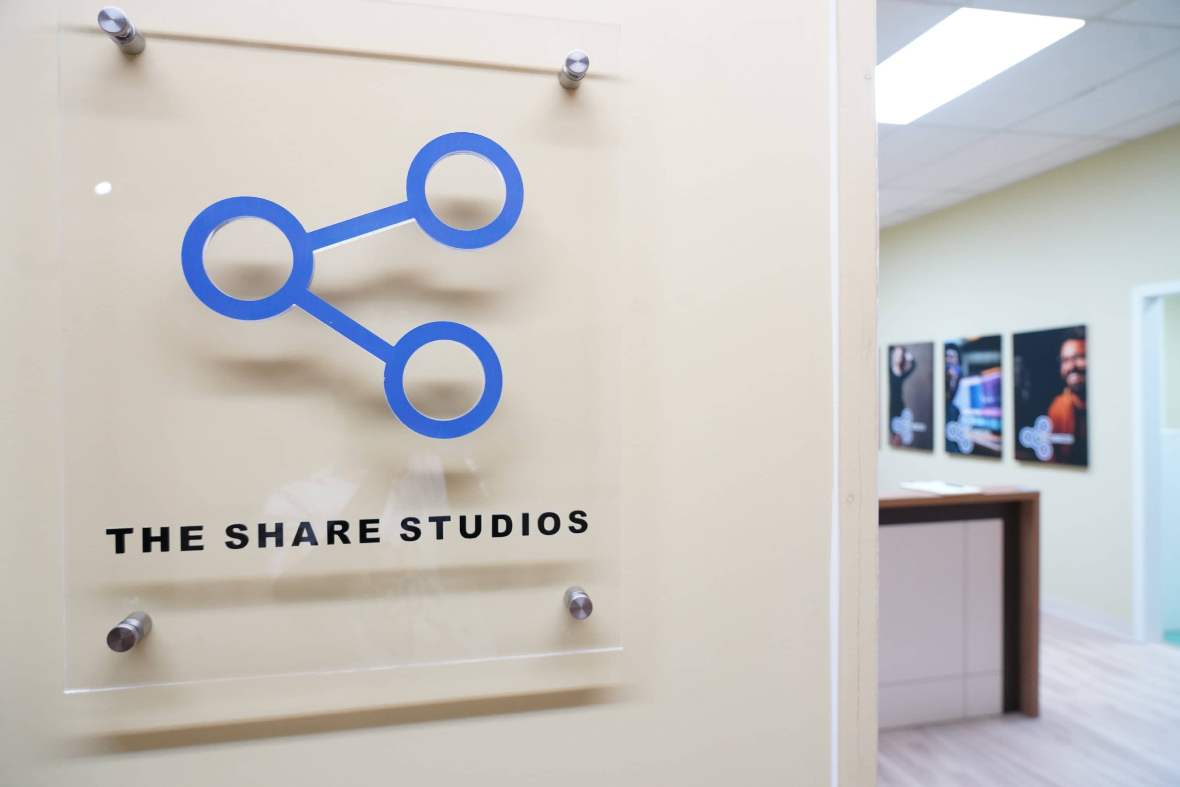 A sign displaying the logo of "The Share Studios" is mounted on a wall near an office interior with framed images visible in the background.