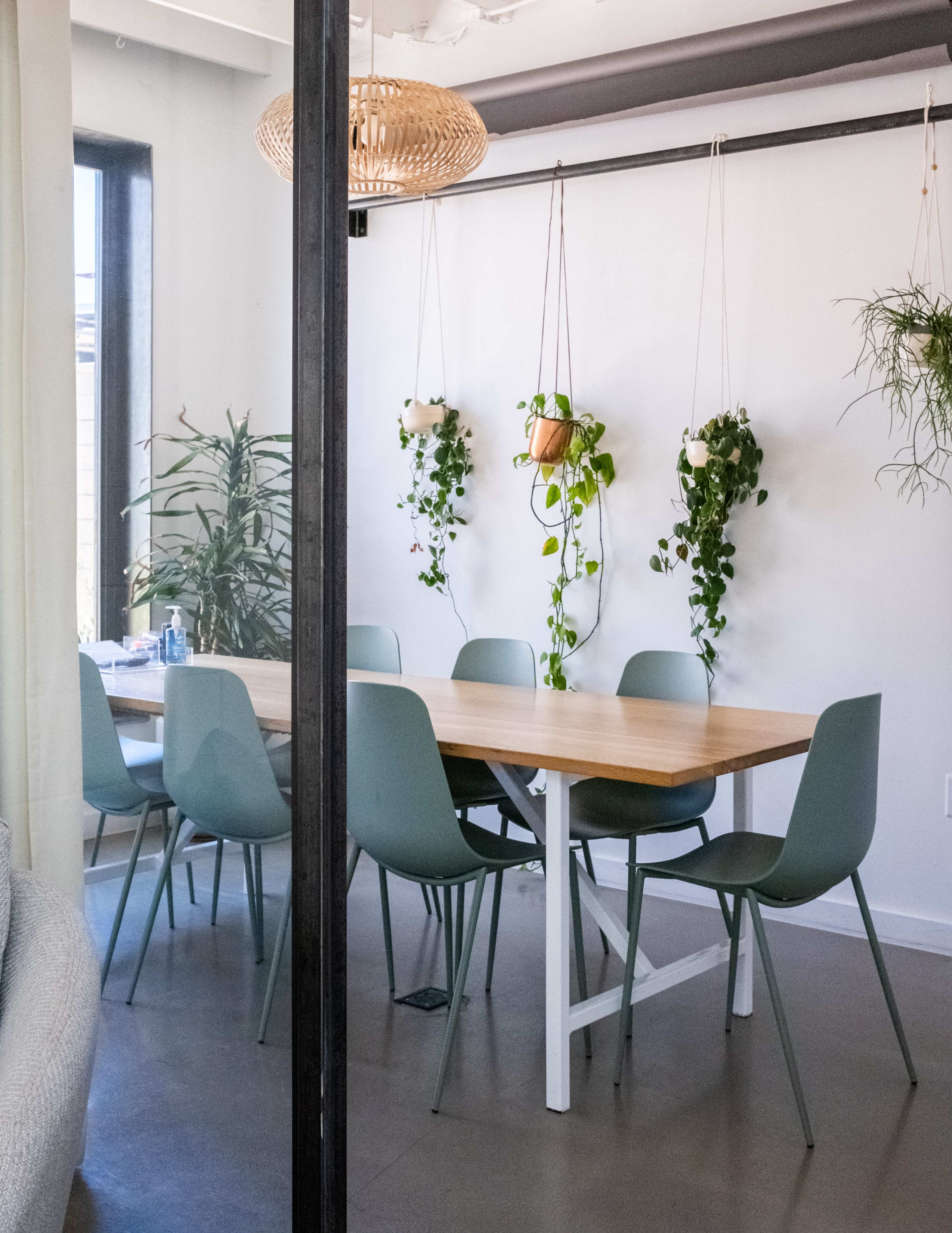A modern dining area features a wooden table surrounded by green chairs, with hanging plant pots and a large window providing natural light.