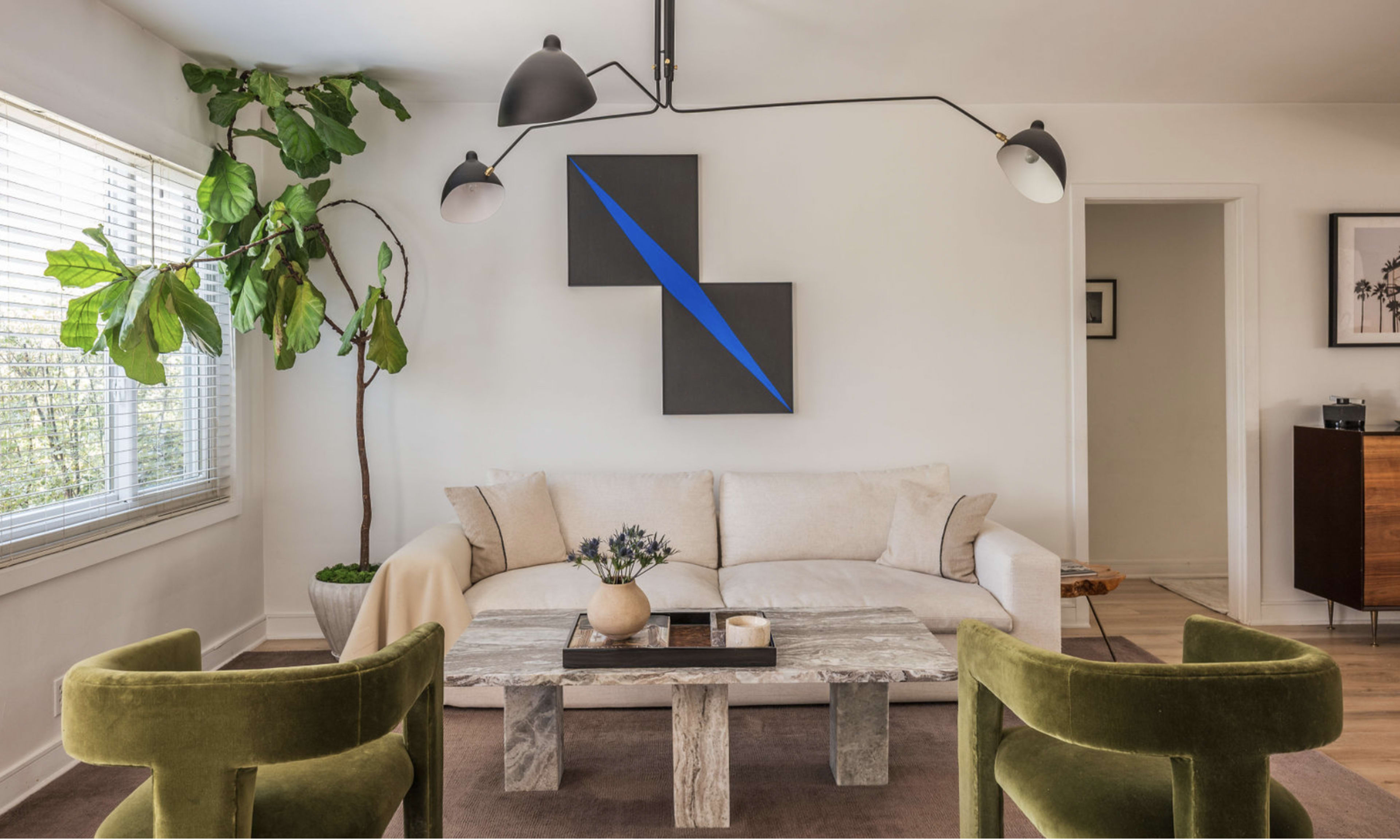 The image depicts a modern, minimalist living room featuring a light-colored sofa, a geometric artwork on the wall, and two green armchairs.