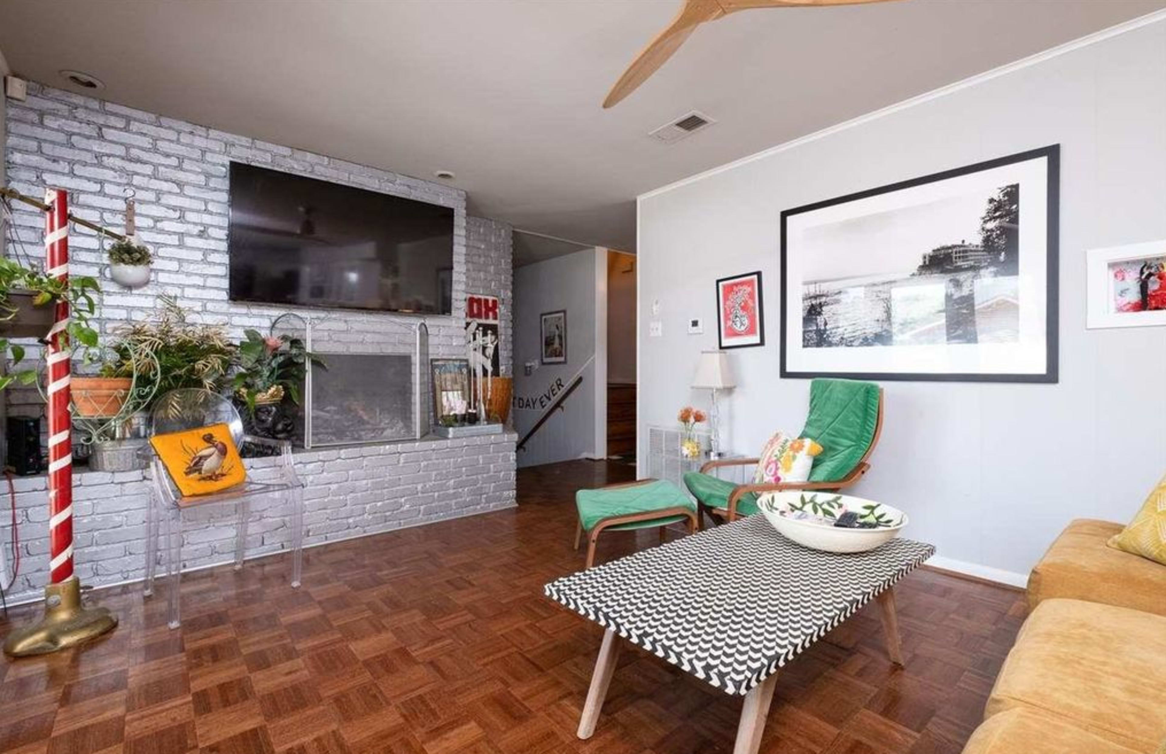 The living room features a brick fireplace surrounded by potted plants, a television mounted above, and seating arranged around a patterned coffee table.
