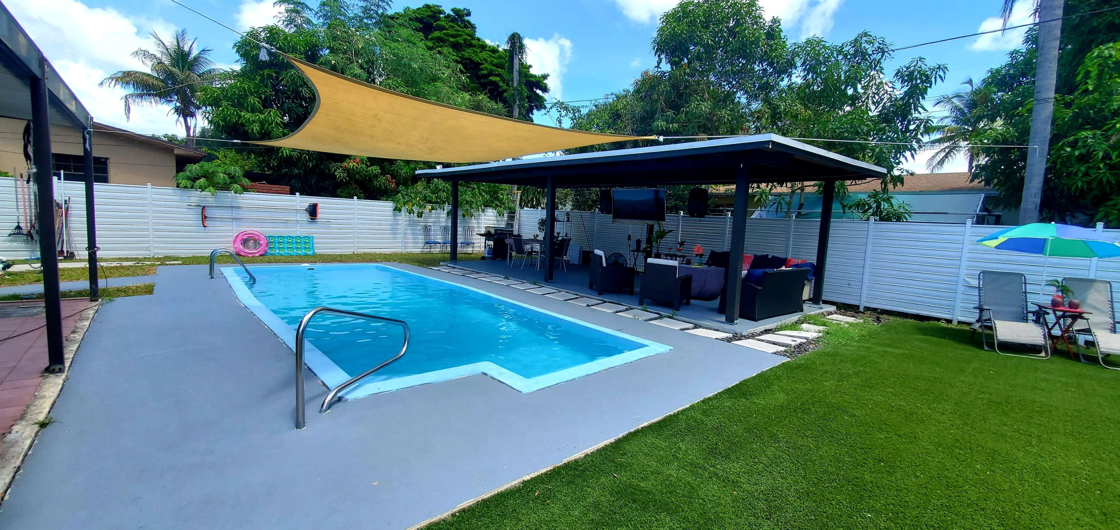 The image shows a backyard pool area featuring a rectangular swimming pool, a shaded seating area with outdoor furniture, and a maintained lawn.