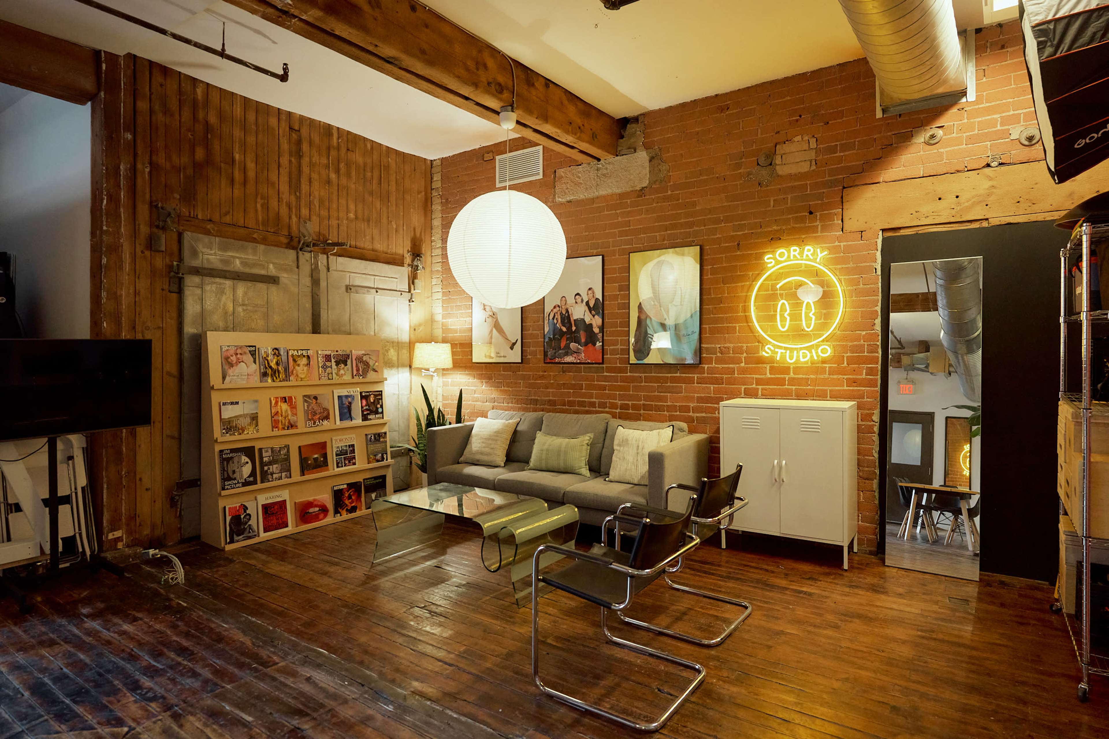 The image shows a cozy studio space featuring brick walls, wooden flooring, a sofa with green cushions, and various artwork, along with a neon sign that reads "SORRY STUDIO."