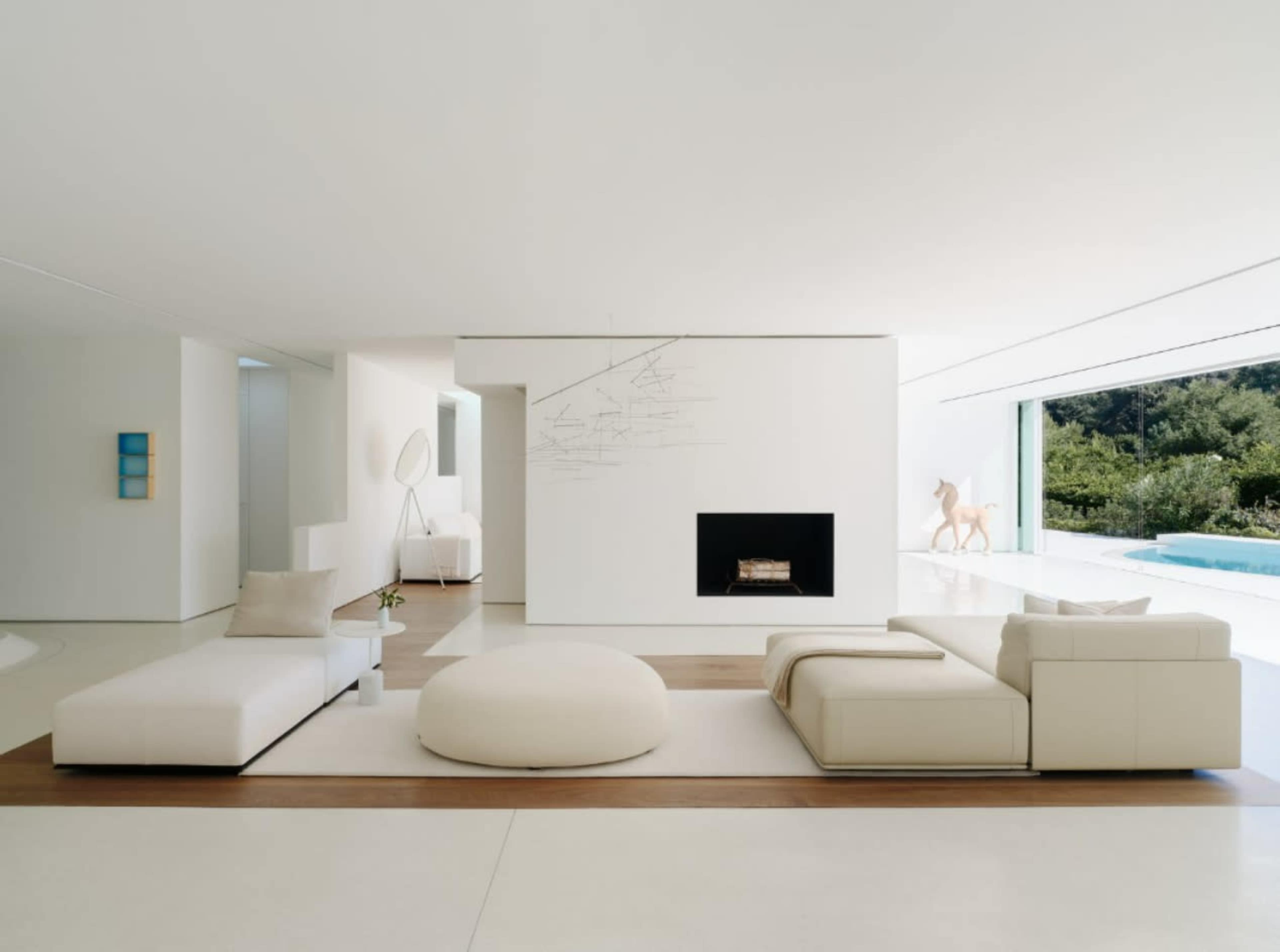 The image shows a modern living room with white walls, minimalist furniture, and a large window overlooking a pool area.
