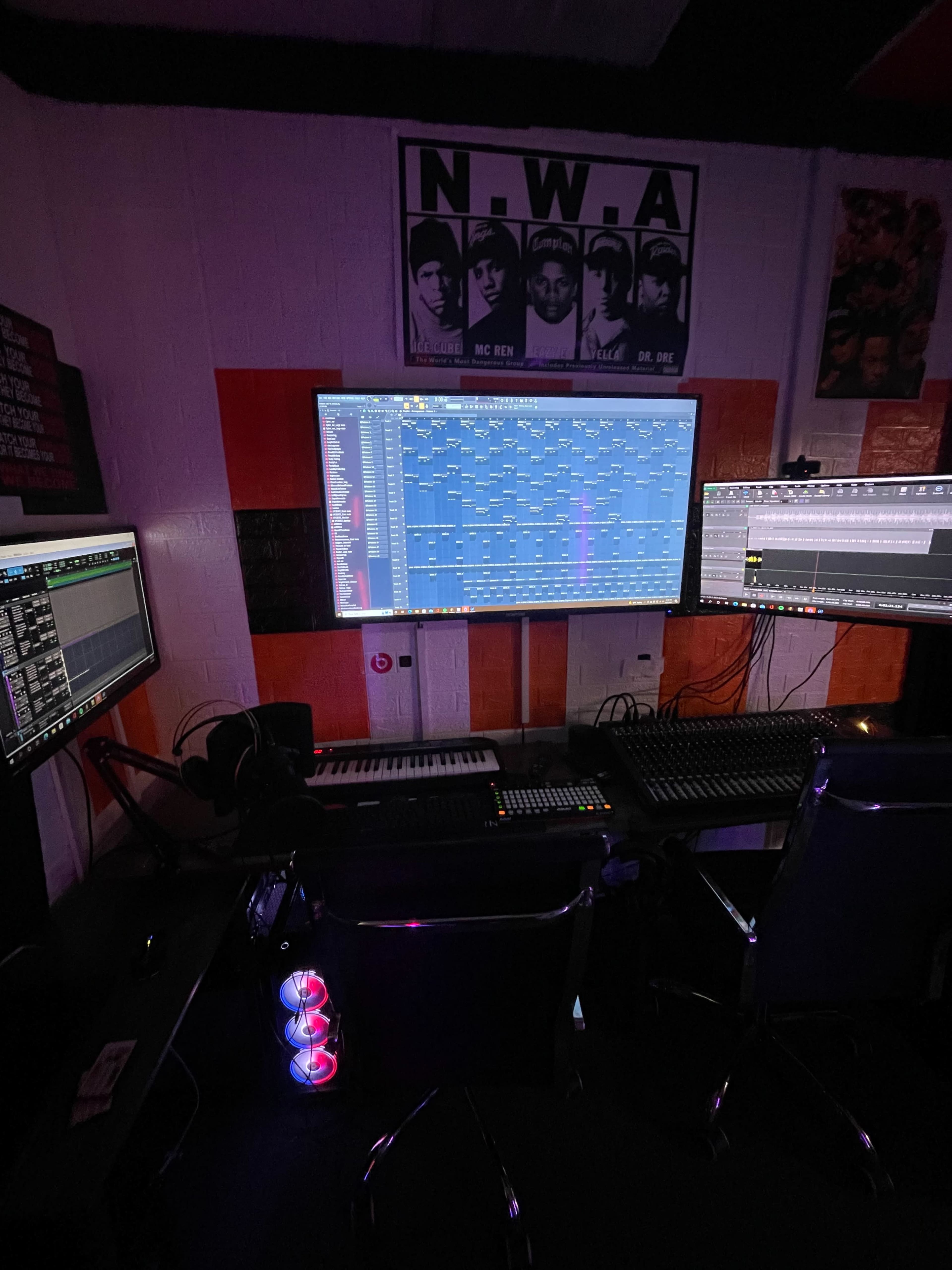 A music production studio with multiple computer monitors displaying digital audio workstations and a colorful LED speaker on the floor.