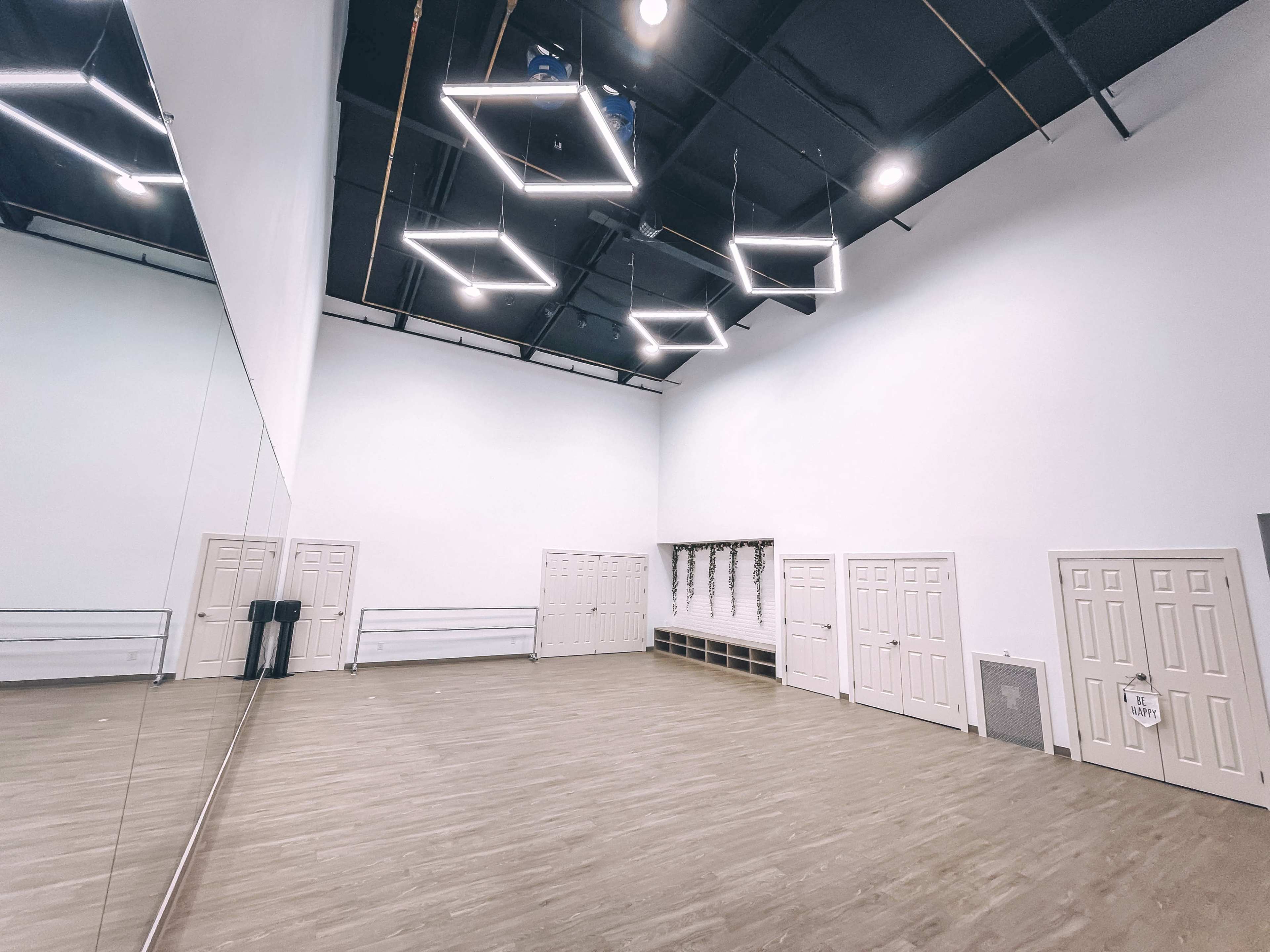 A spacious, empty studio with wooden floors, a large wall mirror, and several doors along one side.