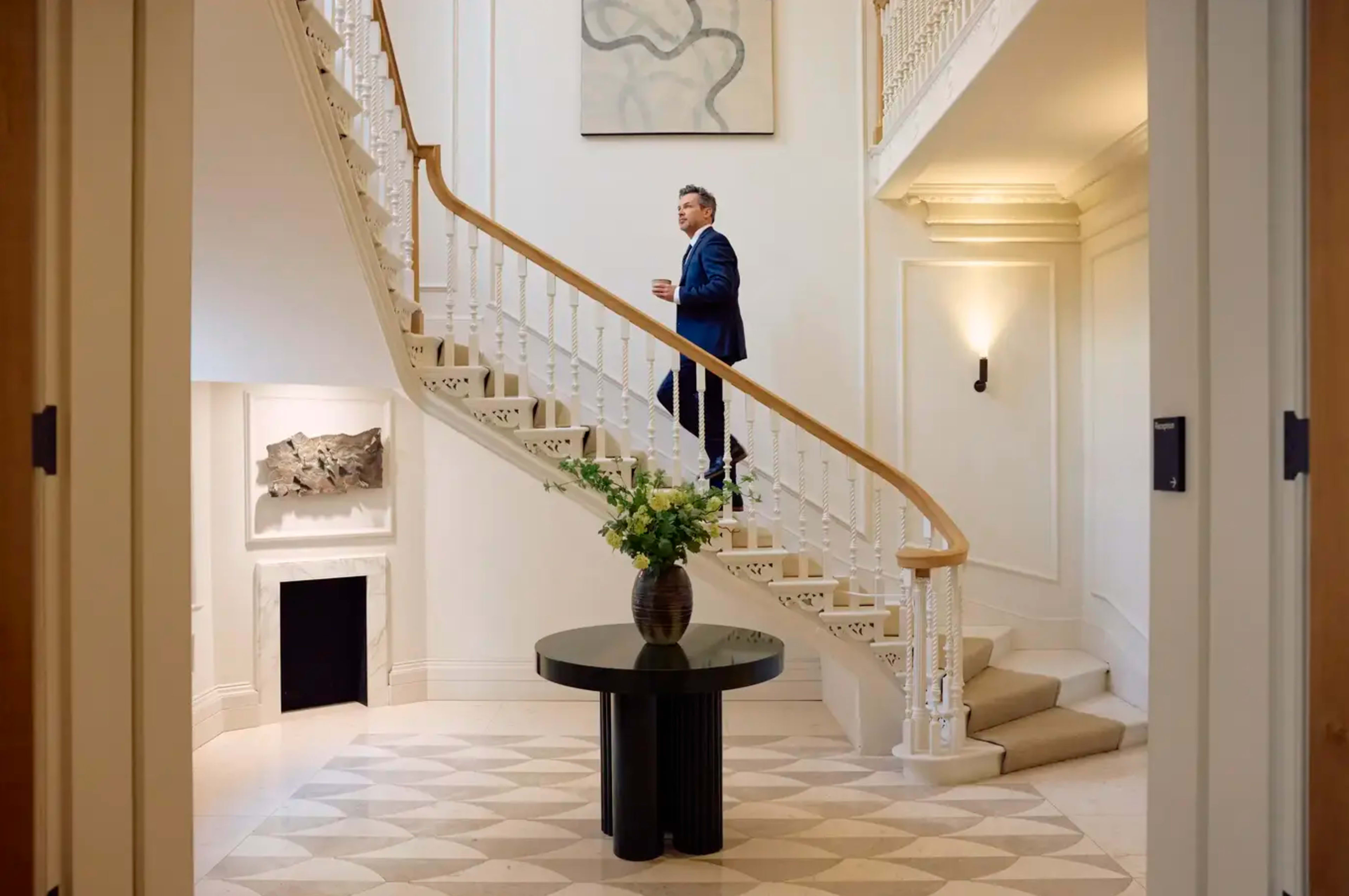 A man in a blue suit is walking up a staircase in a well-lit foyer with a circular table and a flower arrangement.