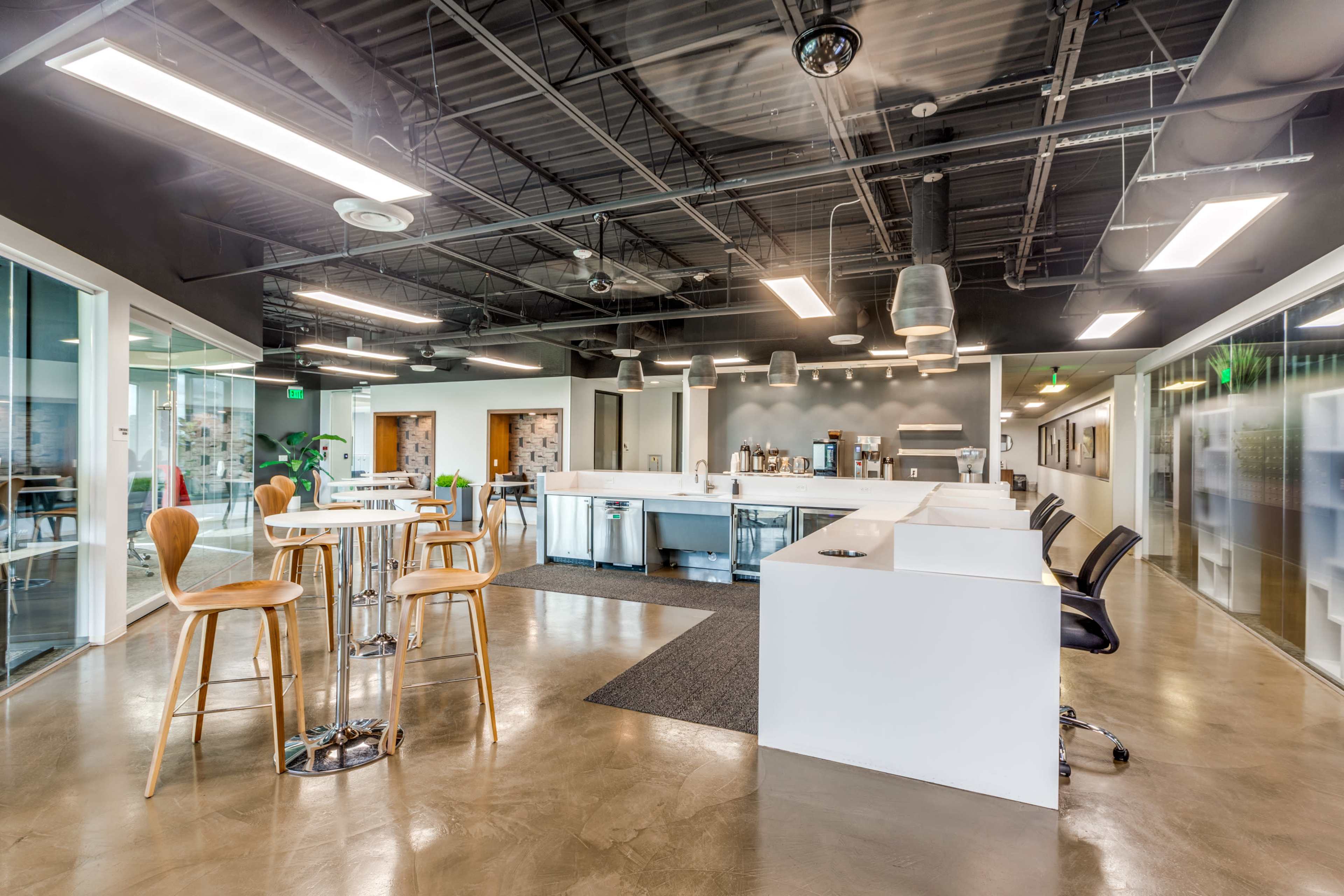 The image shows a modern office break area featuring white cabinetry, seating arrangements with stools and chairs, and large windows allowing natural light.