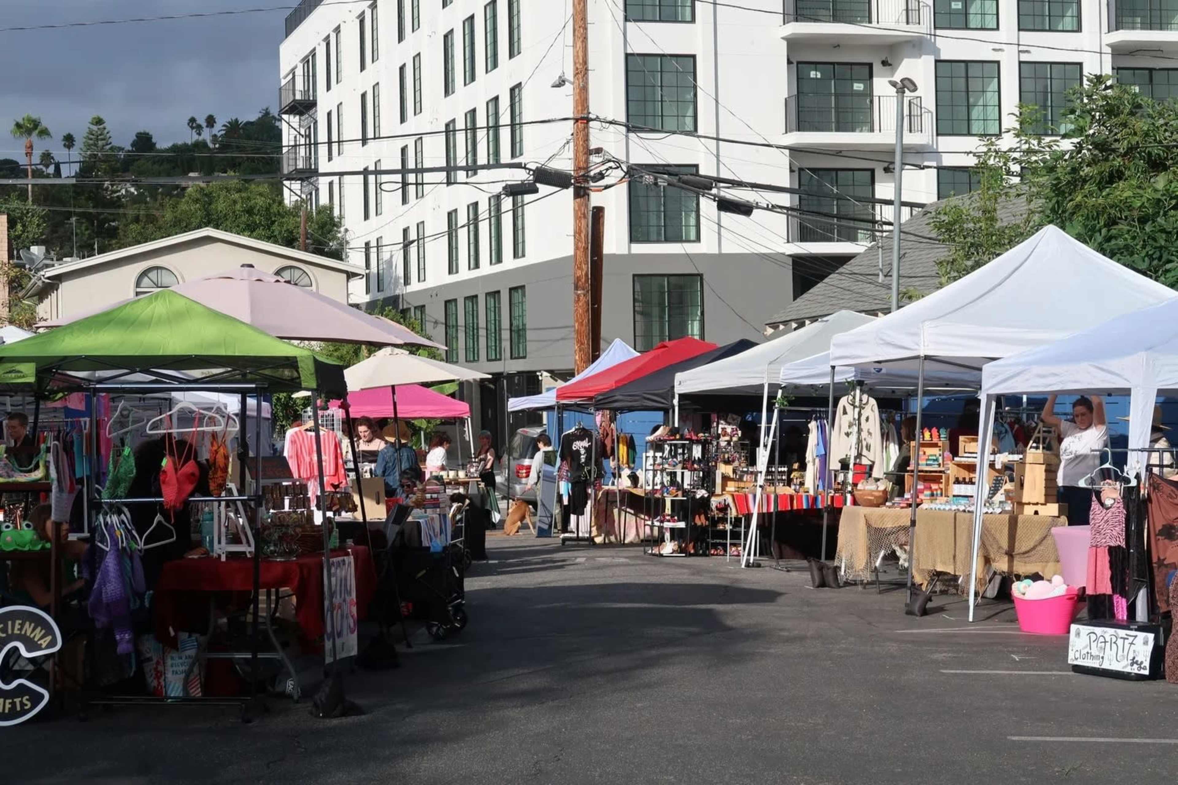 The scene shows a bustling outdoor market with various vendor tents displaying merchandise, set against a backdrop of modern buildings.