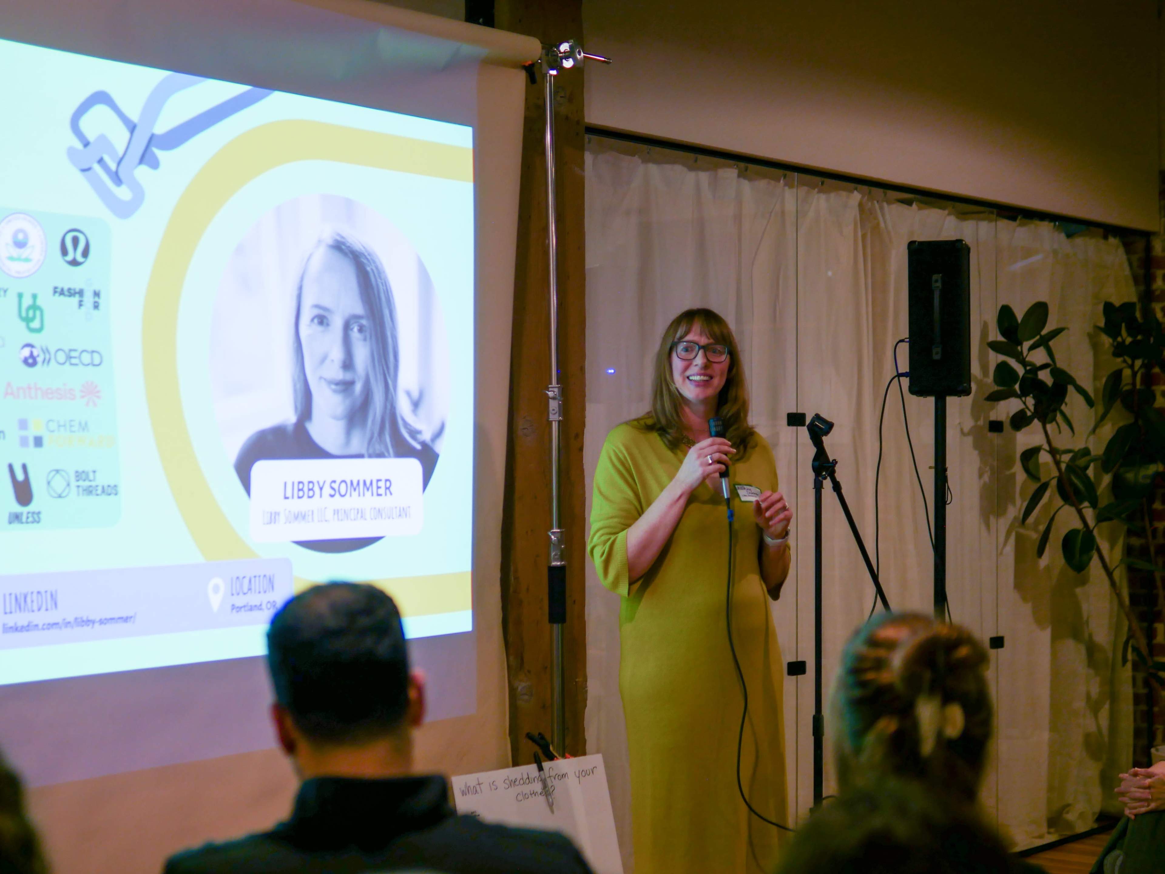 A woman in a green dress speaks at a microphone in front of a projector screen displaying her name and social media links.