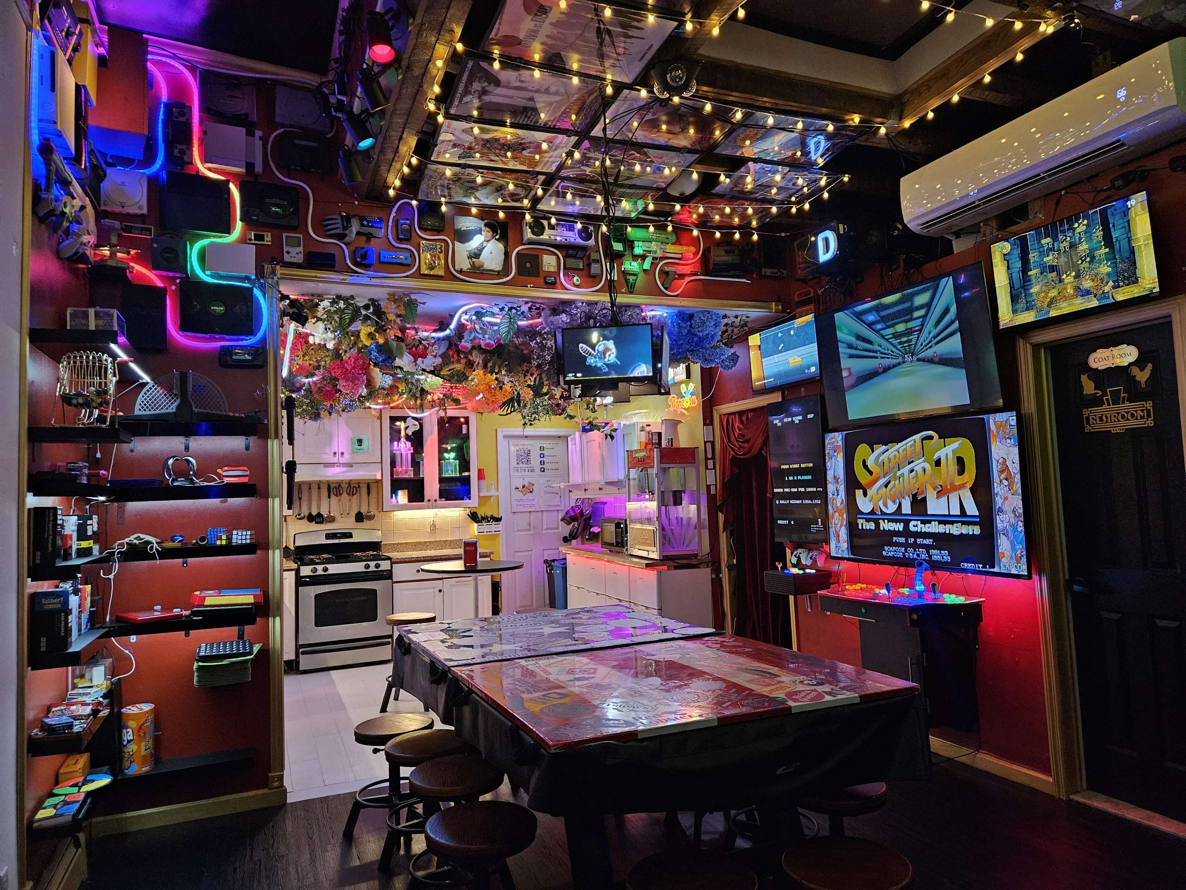 The image shows a colorful gaming room decorated with neon lights, multiple screens displaying games, and a central table surrounded by shelves filled with gaming memorabilia.