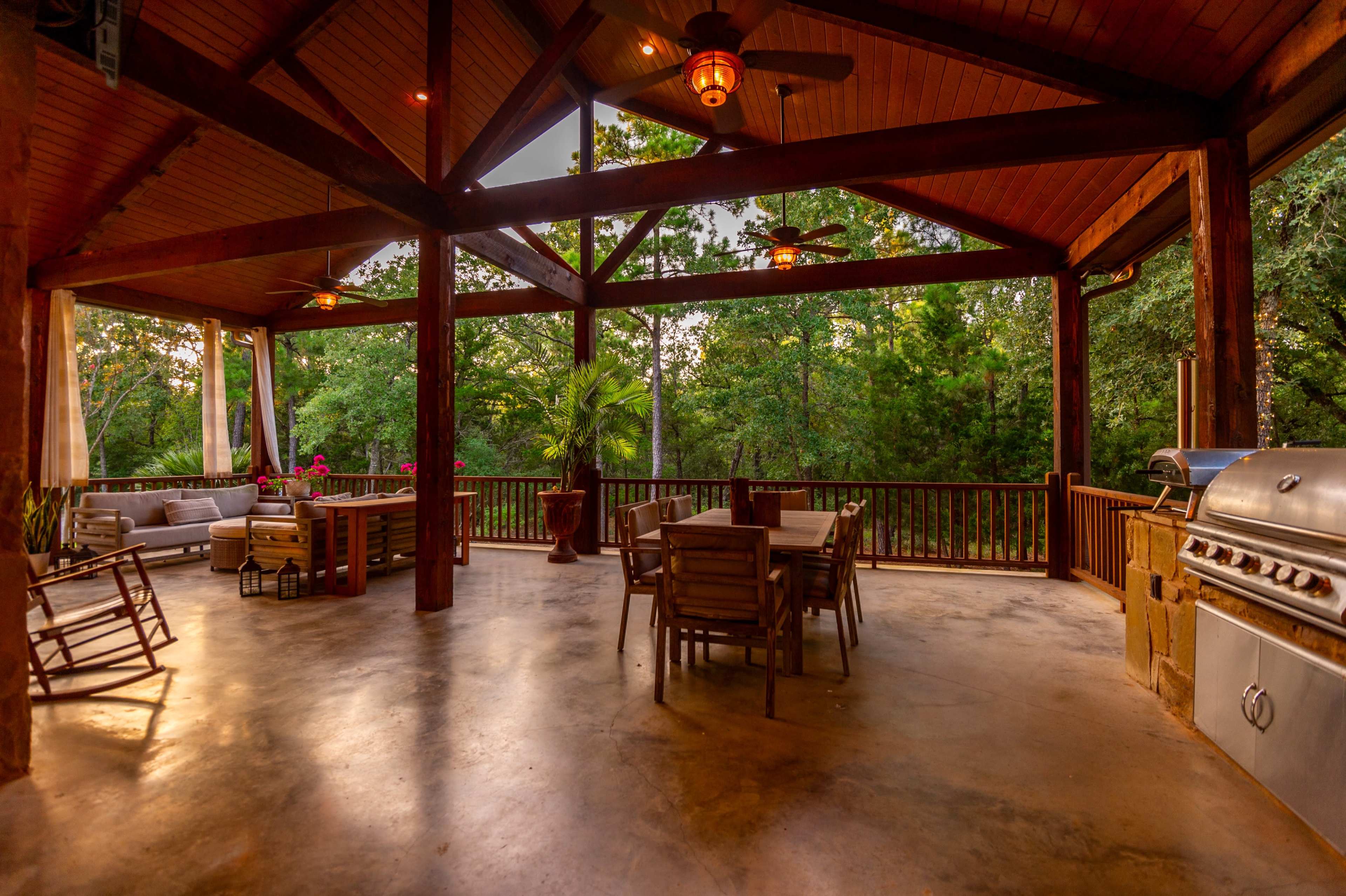 A spacious outdoor patio with wooden beams, a dining area, and a barbecue grill, surrounded by trees.