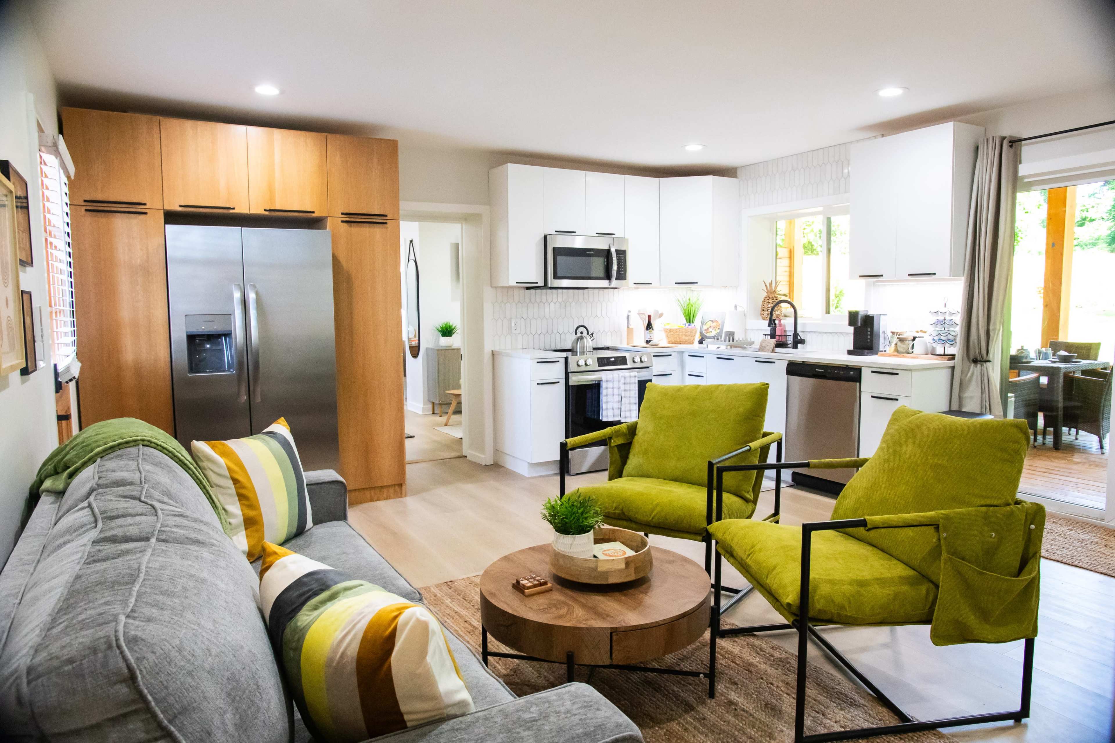 A modern open-concept living space featuring a cozy seating area with two green chairs and a circular wooden coffee table, adjacent to a kitchen equipped with stainless steel appliances and white cabinetry.