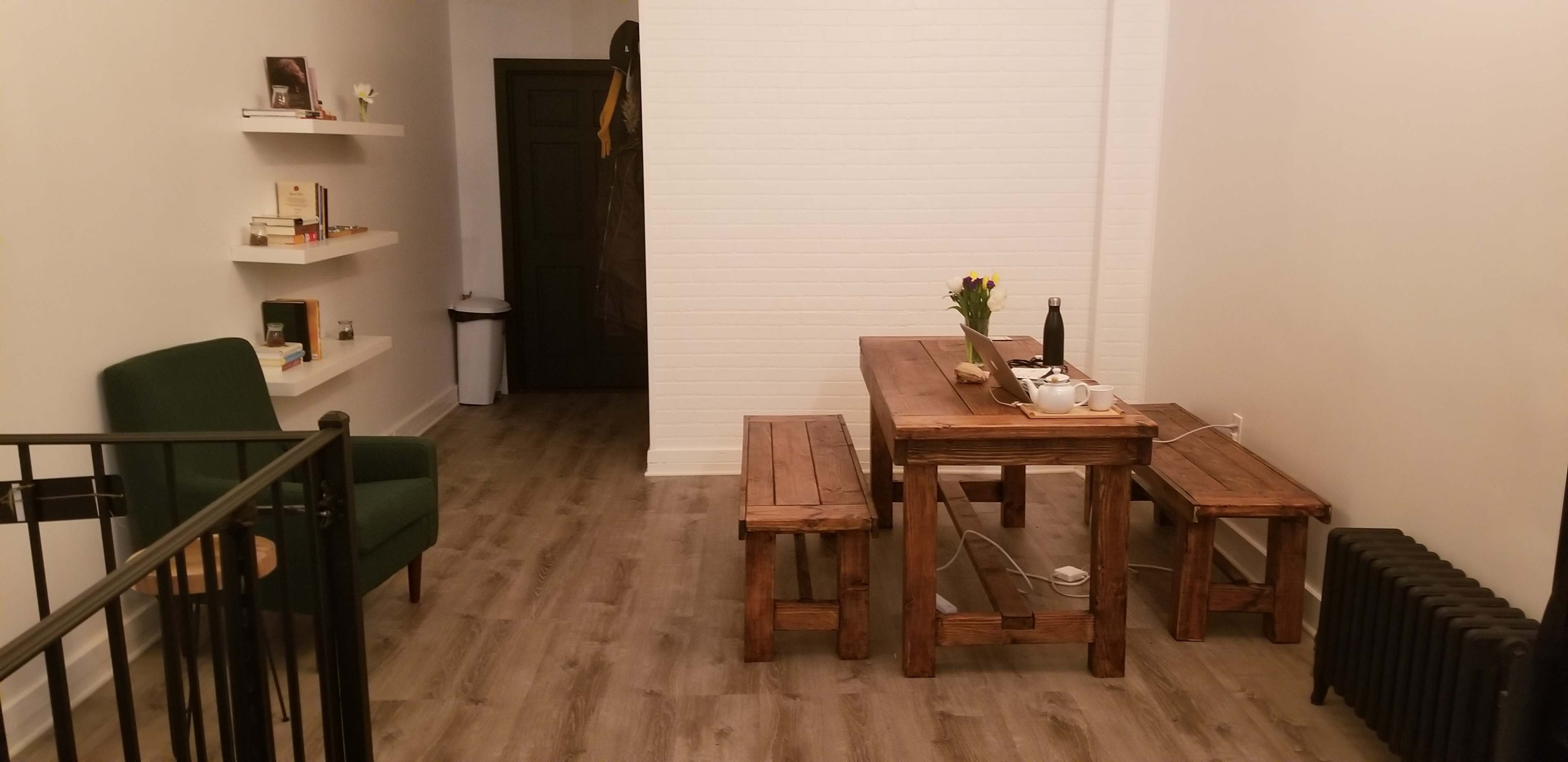 The image shows a simple, minimalist room featuring a wooden table with benches, a green chair, and a shelf with books against a white wall.