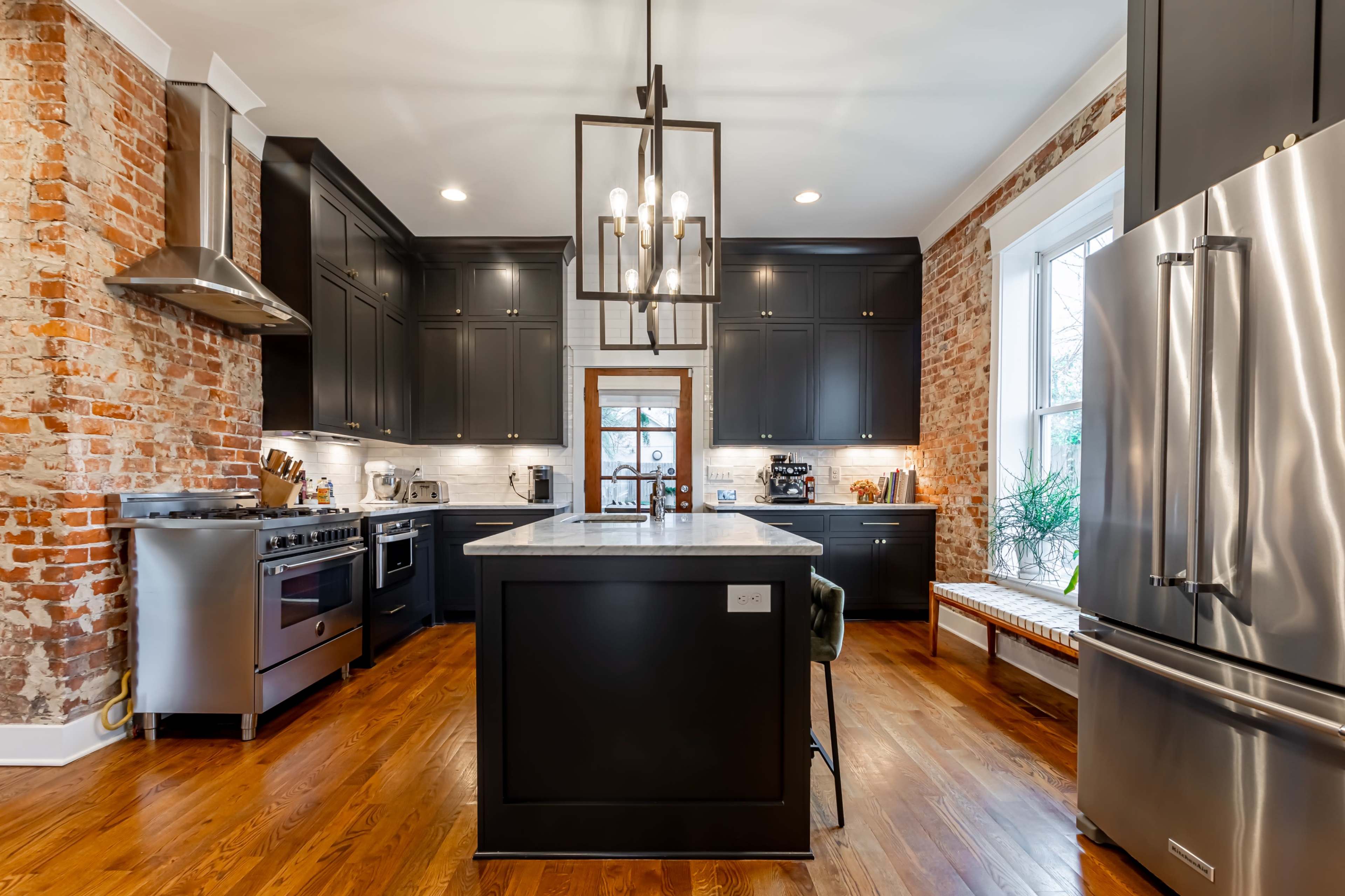 The kitchen features exposed brick walls, dark cabinetry, a marble island, and stainless steel appliances.