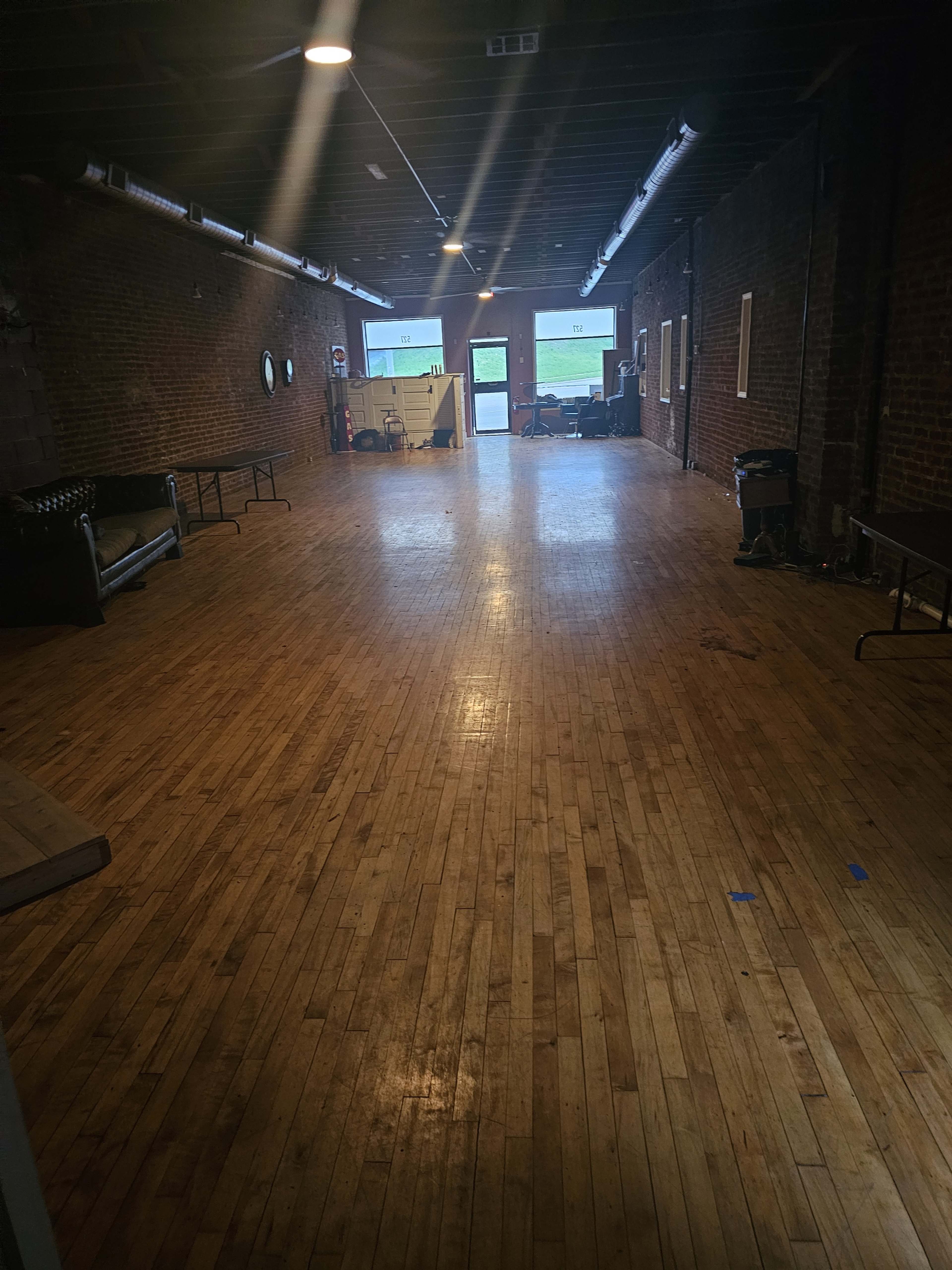 A spacious, empty room with wooden flooring, exposed brick walls, and large windows at one end.
