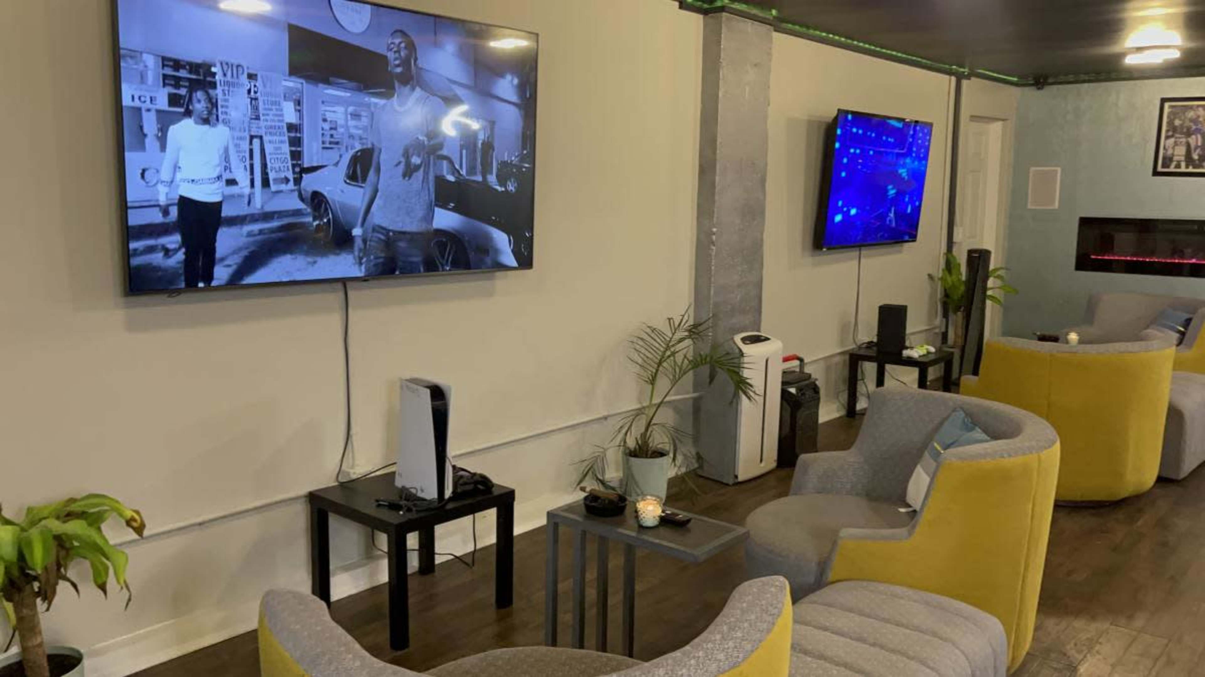 The image shows a cozy lounge area with gray and yellow armchairs, two large televisions mounted on the walls displaying different content, and decorative plants in the space.
