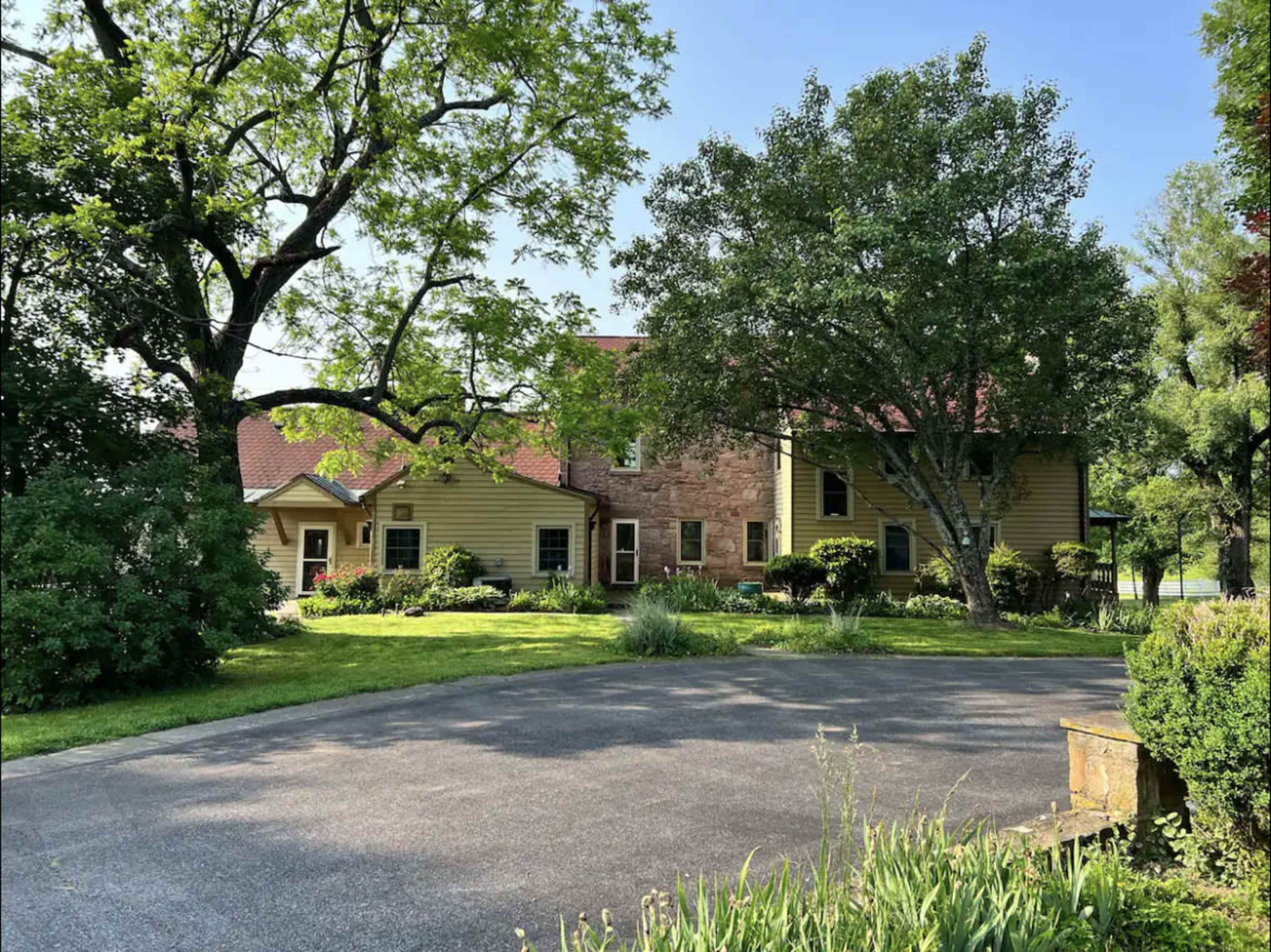 A large house with a mix of yellow siding and stone exterior, surrounded by trees and a well-maintained lawn, with a paved driveway leading up to it.