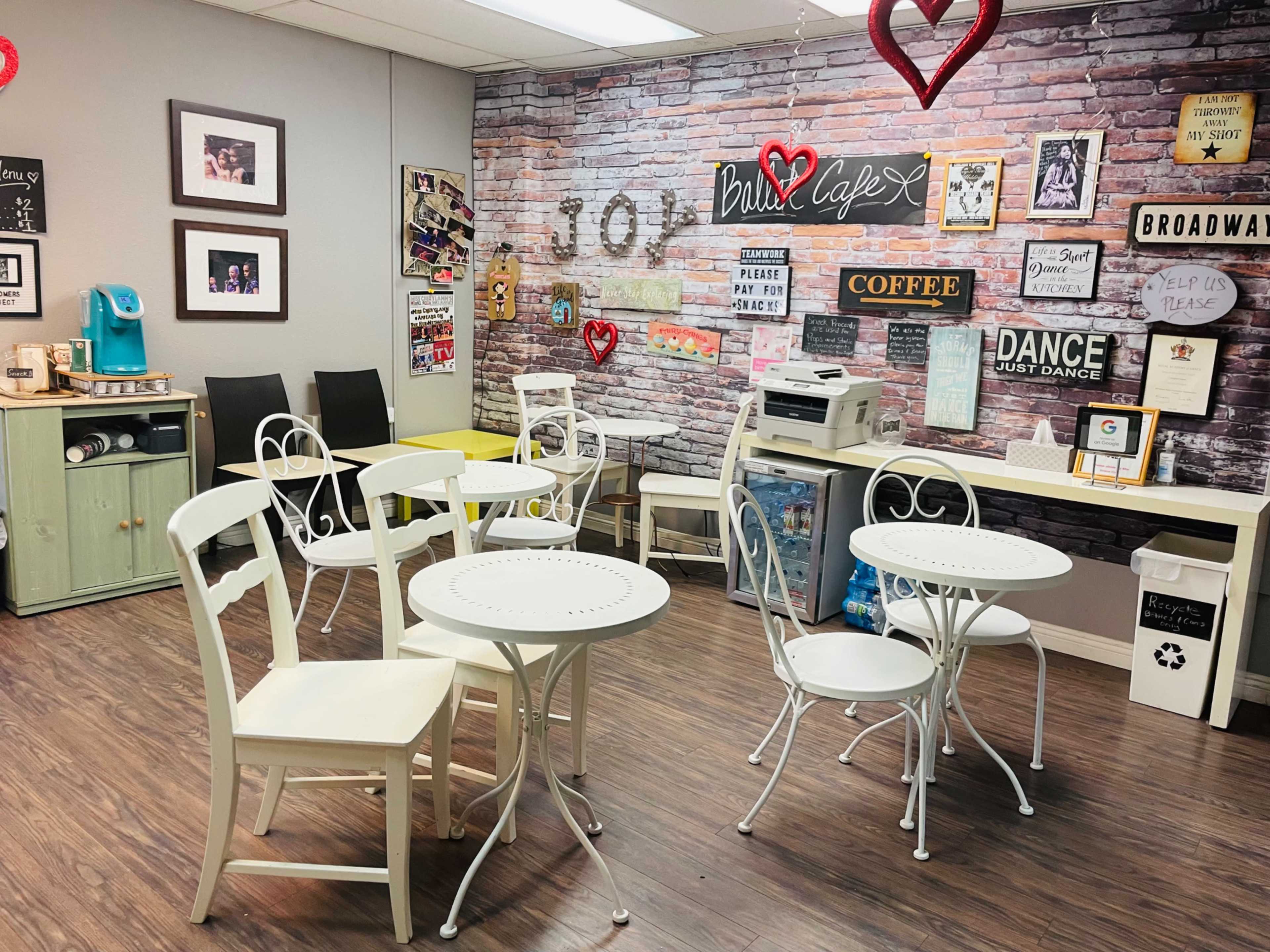 A quaint café interior with white metal tables and chairs, a printer, and a wall decorated with posters and a brick design.