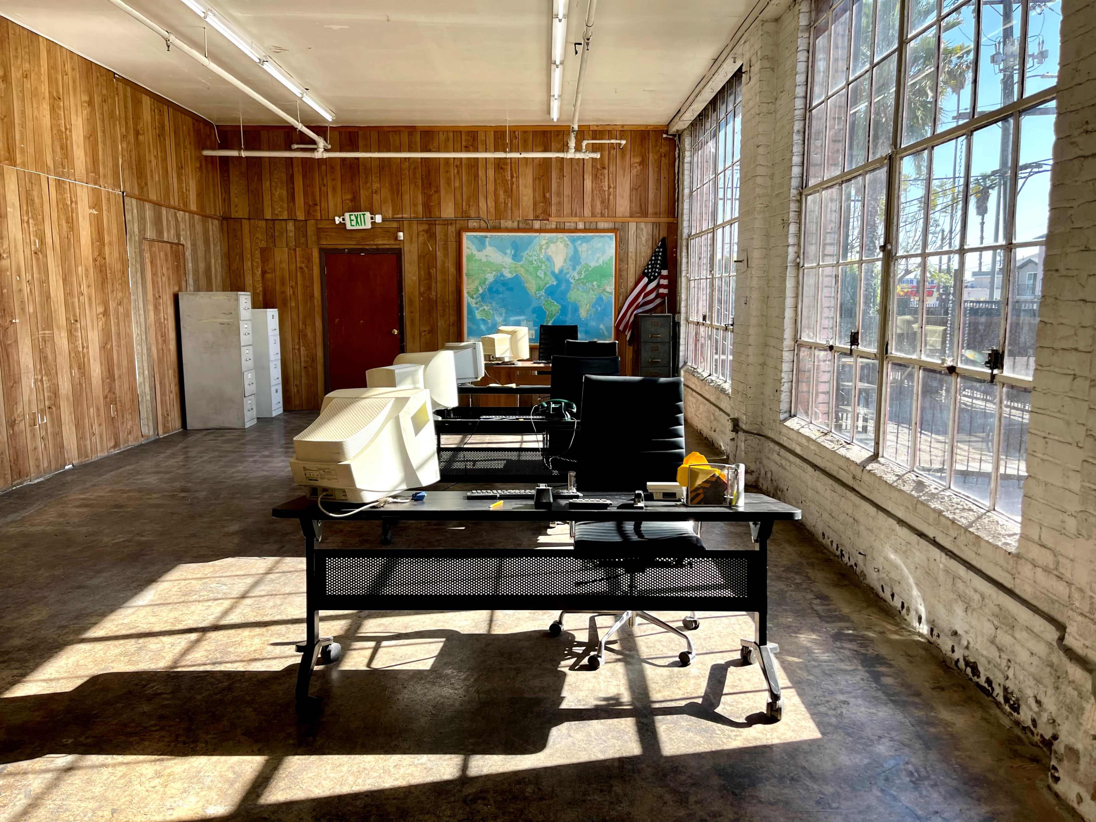 The image shows an empty office space with several vintage computers and chairs arranged at desks near large windows, featuring a map on the wall and wooden paneling.